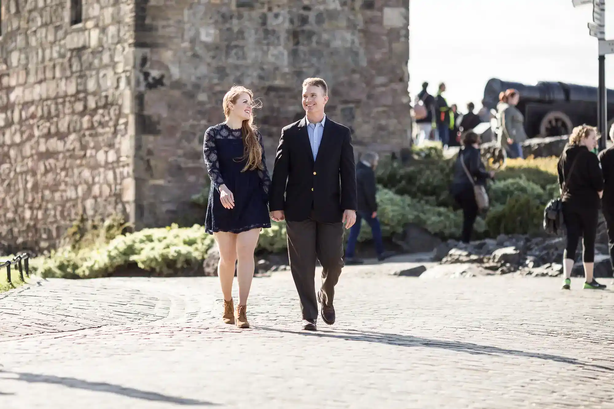 A smiling couple walk hand in hand on a sunny cobbled path near an old stone building, with other people and a cannon blurred in the background.