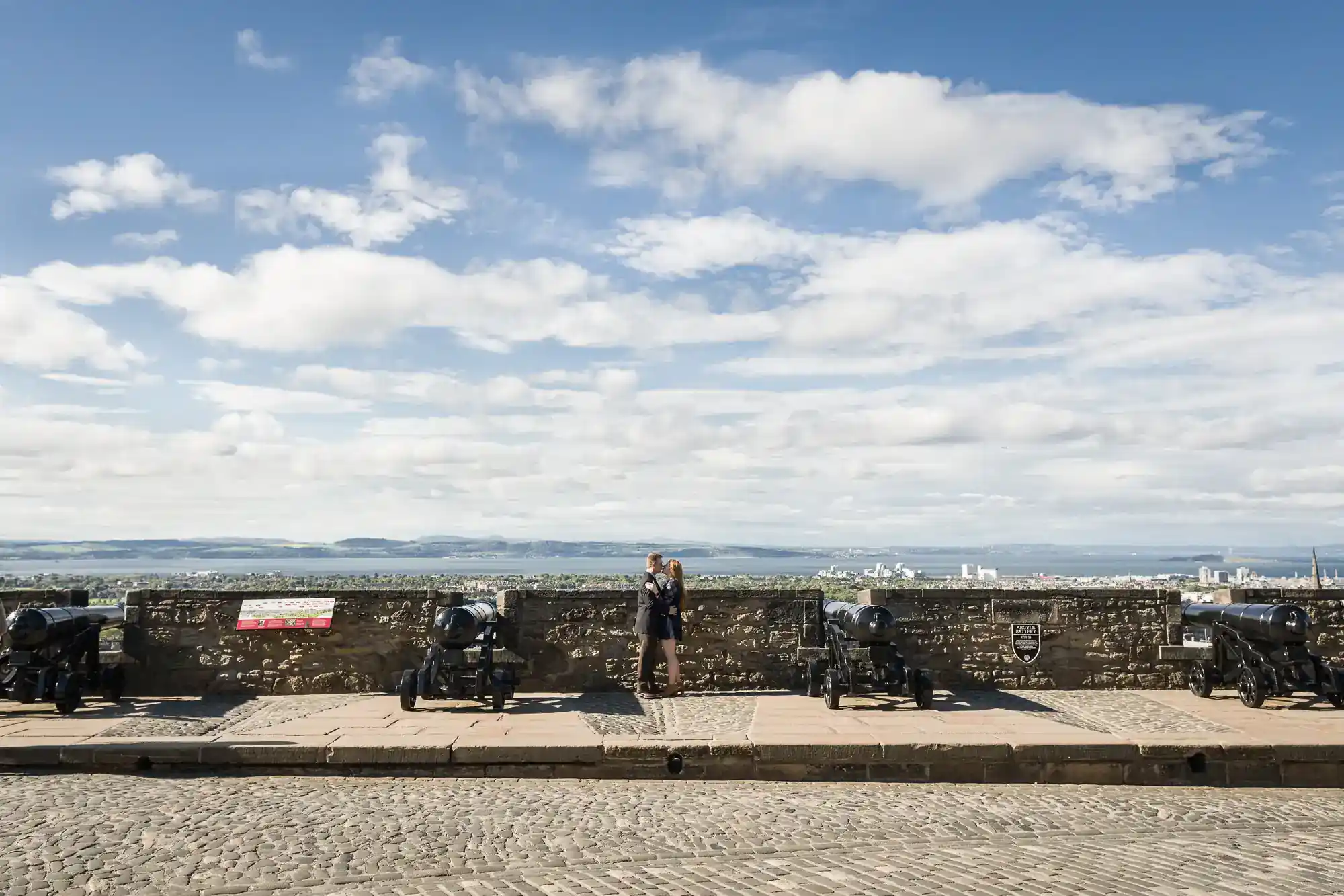 Five black cannons are lined up along a stone wall overlooking a distant cityscape and mountains under a blue sky with scattered clouds. A person stands at the centre, looking out over the view. The foreground has cobblestone pavement.