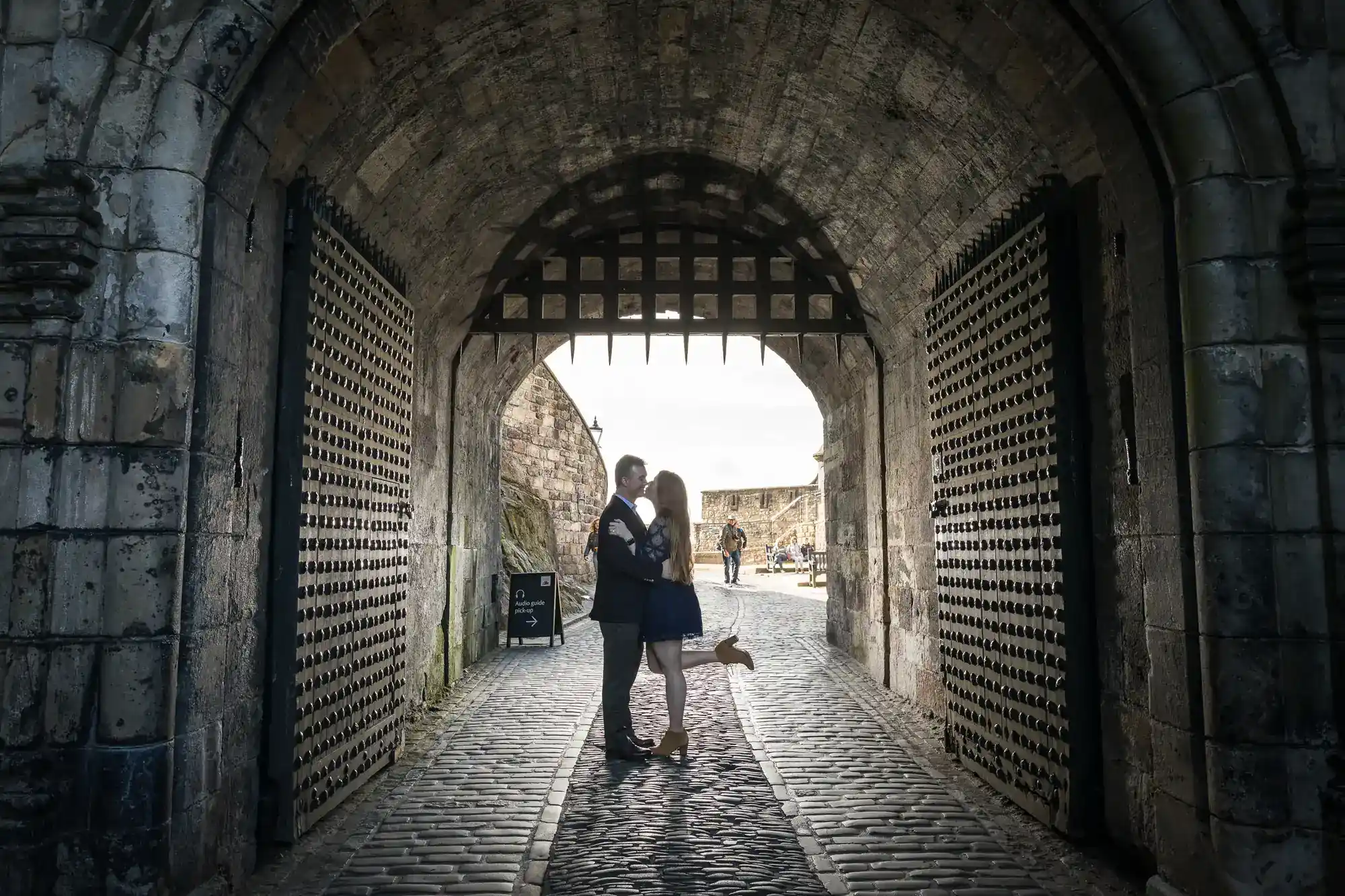 A couple embraces under a stone archway with an iron gate above, standing on cobblestones. The woman lifts one leg playfully. Sunlight streams from the open end of the tunnel. Large studded doors frame the scene. A person is visible in the background.