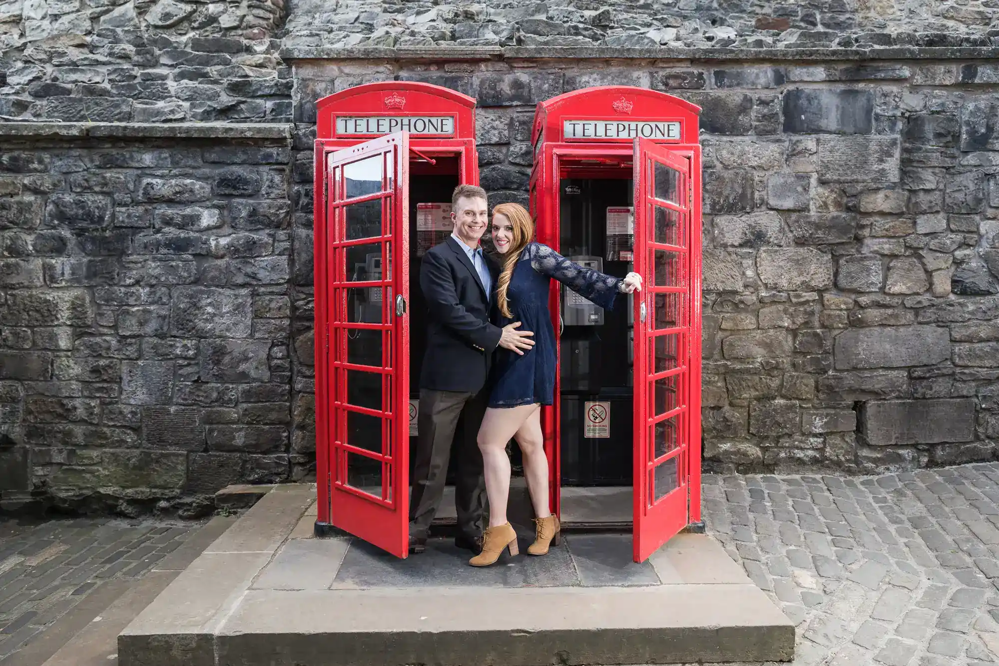 A smiling couple stands together, posing between two classic red British phone boxes in front of a weathered stone wall. The woman wears a short dress and boots, hugging the man, who is dressed in a suit. Both doors of the boxes are open.