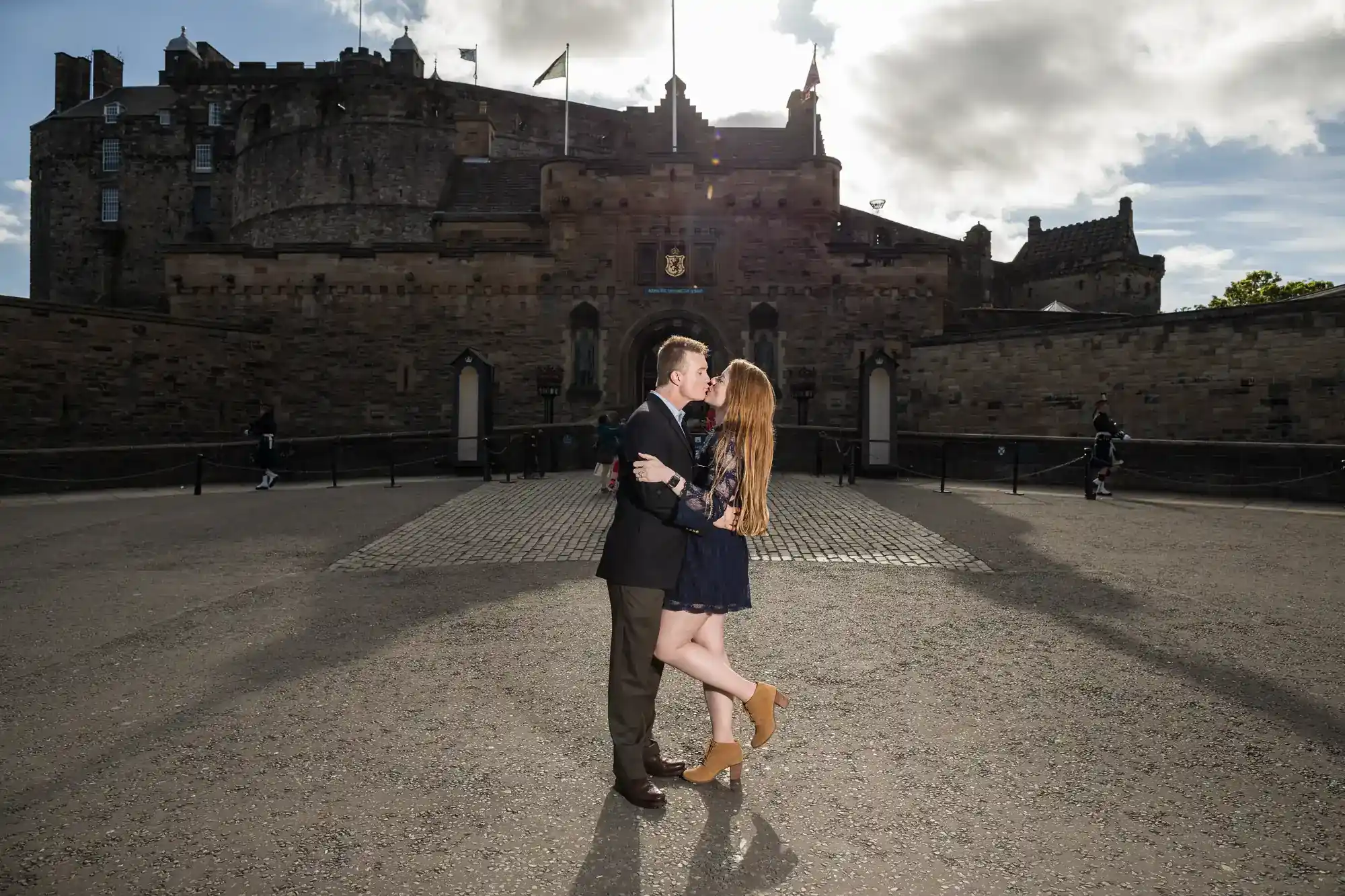 A couple embraces and kisses in the centre of a paved courtyard in front of a historic stone castle with towers and flags under a cloudy sky, sunlight shining behind the castle.