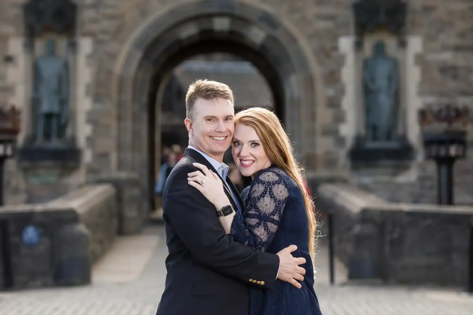 A smiling couple embrace in front of a castle entrance. The man wears a dark suit; the woman has long red hair and a navy lace dress. Stone statues and arches frame the background, with cobbled paving underfoot, looking towards their Edinburgh Castle wedding photographer.