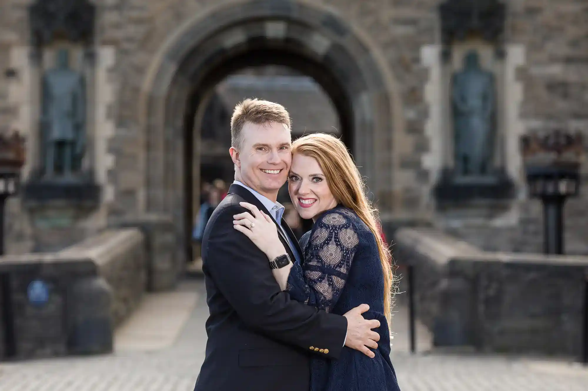 A smiling couple embrace in front of a castle entrance. The man wears a dark suit; the woman has long red hair and a navy lace dress. Stone statues and arches frame the background, with cobbled paving underfoot.
