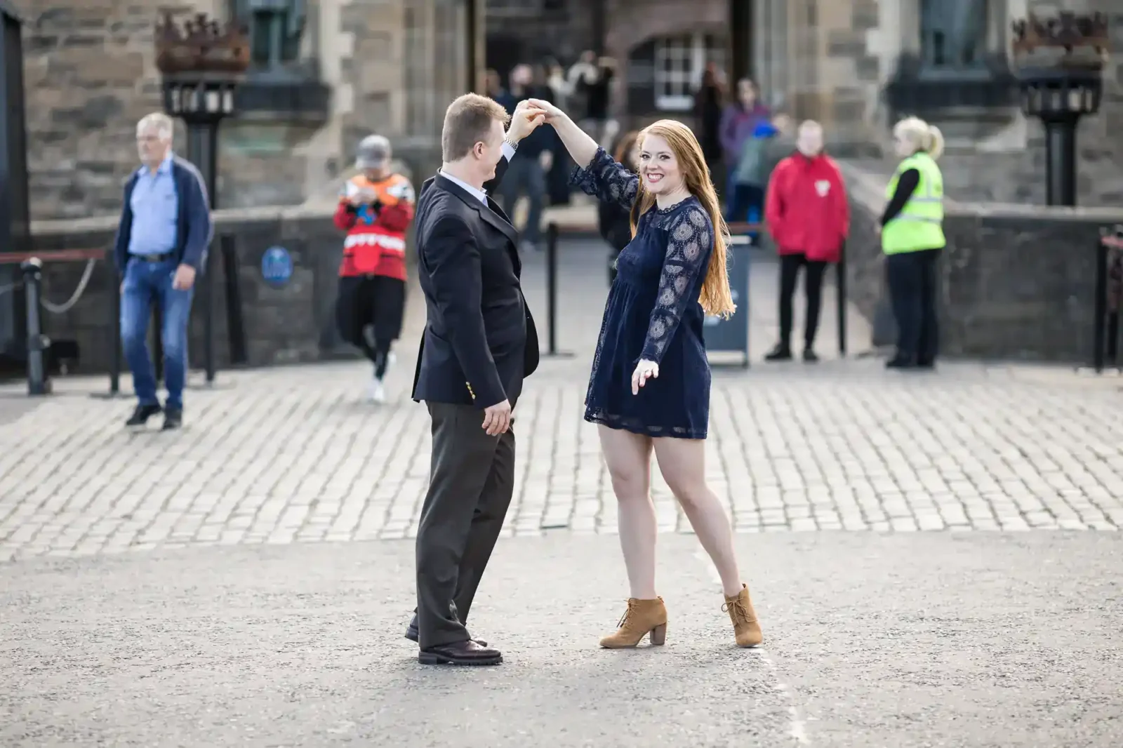 A man in a dark blazer twirls a smiling woman in a navy blue dress and tan boots on a cobbled street, The Esplanade at Edinburgh Castle, with historic stone buildings and several people watching in the blurred background.