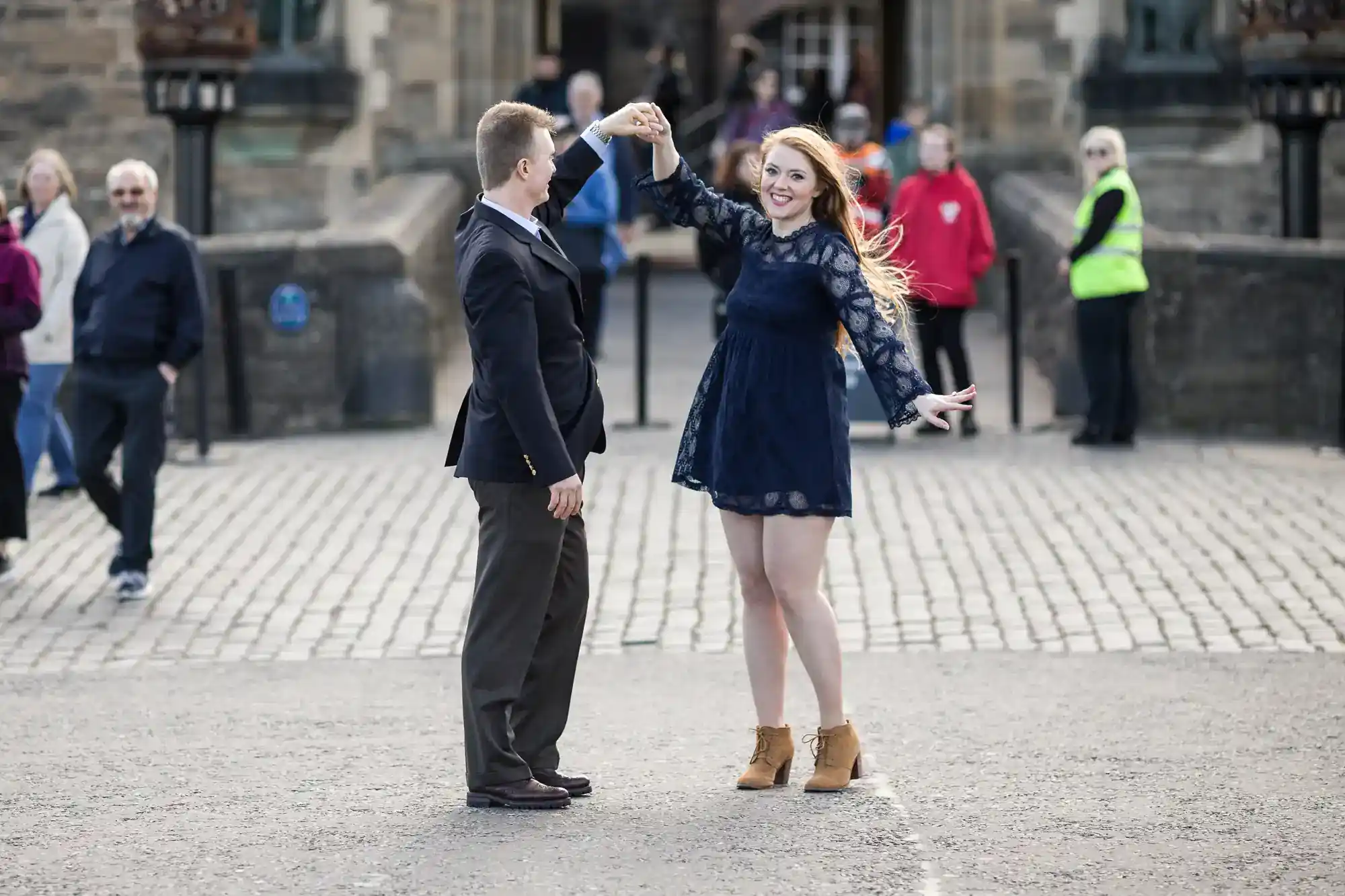 A man in a dark suit twirls a smiling woman in a short navy dress and tan boots in a cobbled square. She holds his hand, looking happy, with people walking and a large stone building visible in the background.