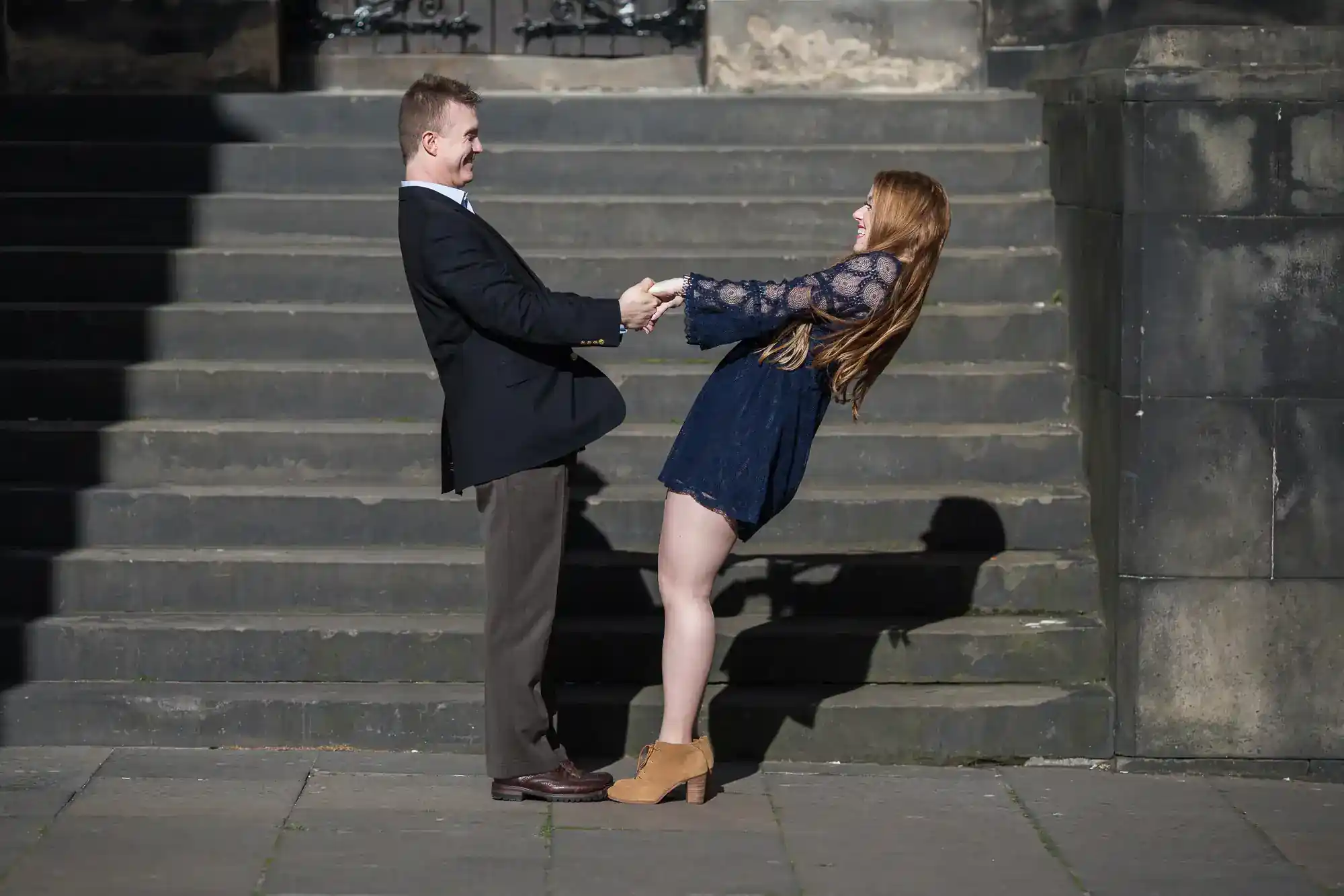 A man in a dark suit and a woman in a short blue dress smiling and holding hands, leaning backwards while facing each other, stand on a stone pavement in front of wide concrete steps on a sunny day.
