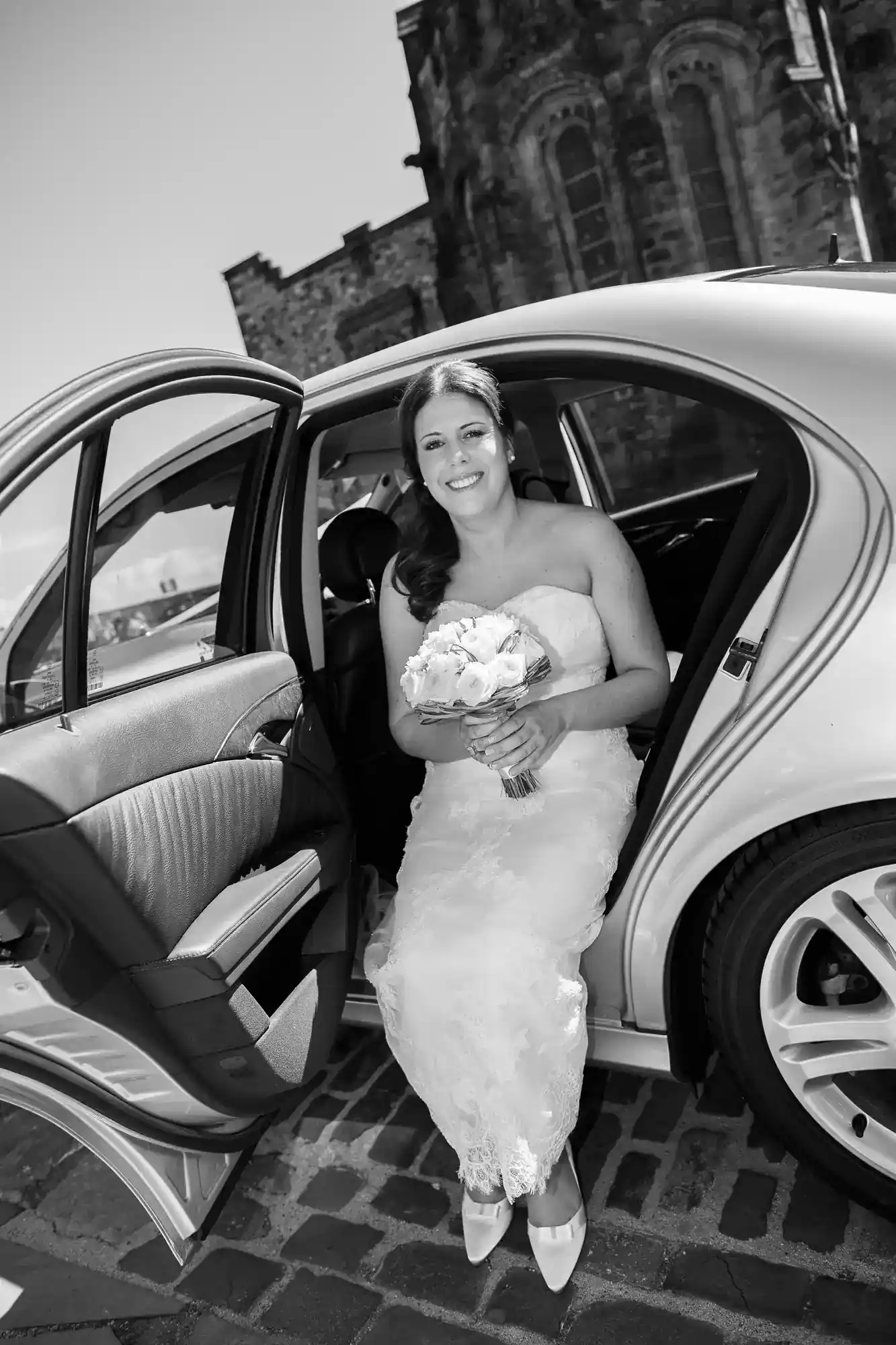 A bride in a strapless white gown sits in the open doorway of a car, holding a bouquet of roses and smiling. The background features an historic stone building. The photo is black and white, and the bride’s shoes are visible on cobbled pavement.