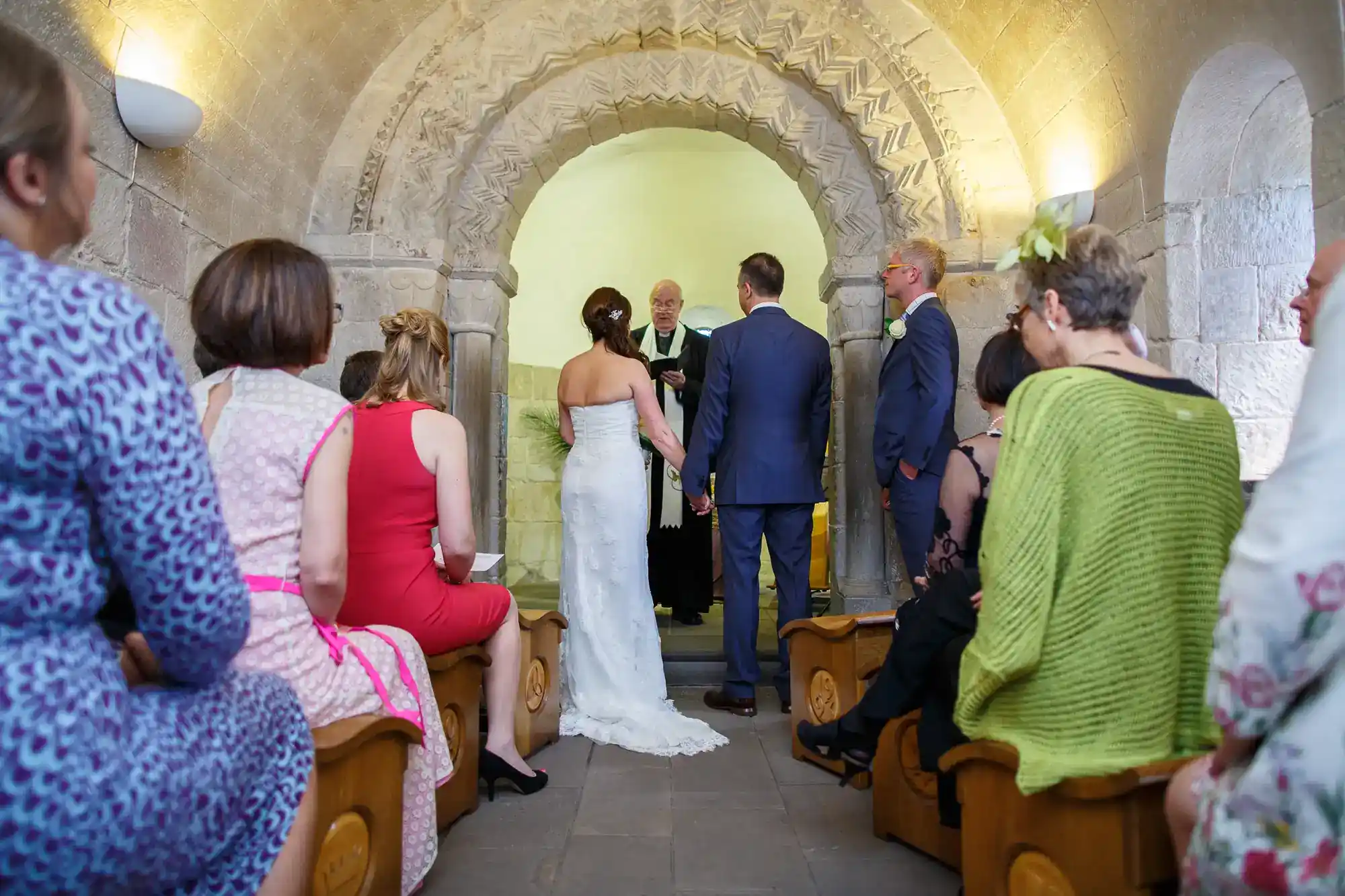 A bride and groom stand at an altar with an officiant in a stone chapel. Guests in colourful attire sit on wooden pews, facing the couple. The arched doorway is ornately carved, and the stone walls are softly lit, creating a warm, intimate setting.