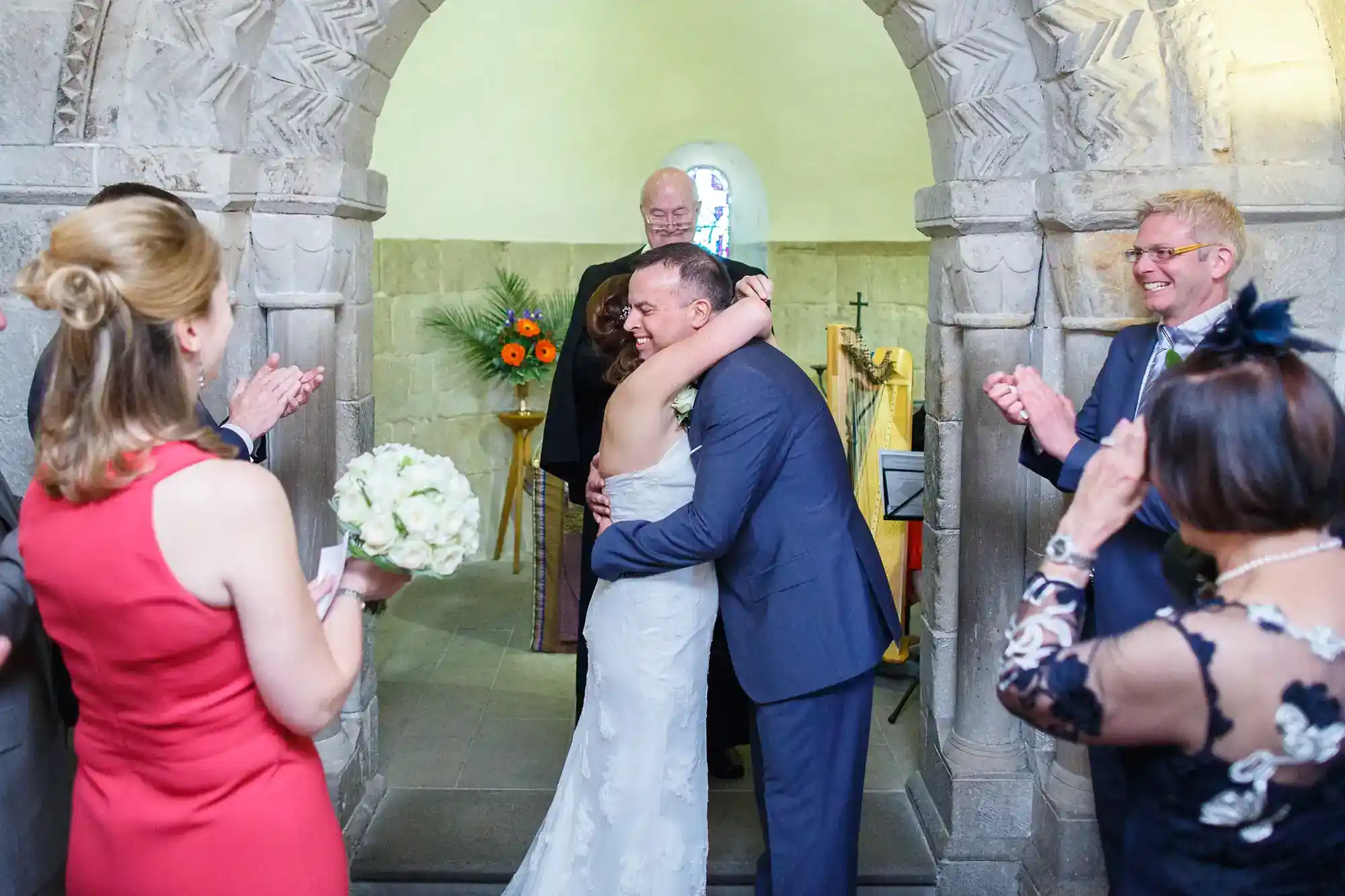 A bride and groom embrace and smile at the altar inside a stone chapel, surrounded by applauding guests. The bride wears a white dress and holds a bouquet, while the groom wears a blue suit. An officiant stands behind them, and flowers decorate the background.