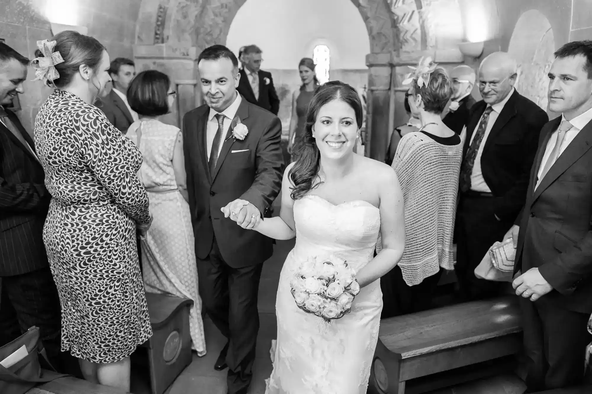 A smiling bride in a strapless gown holds a bouquet, walking down the aisle with a suited man. Guests in formal attire look on, some seated and some standing, in a church with decorated stone walls and arched windows.