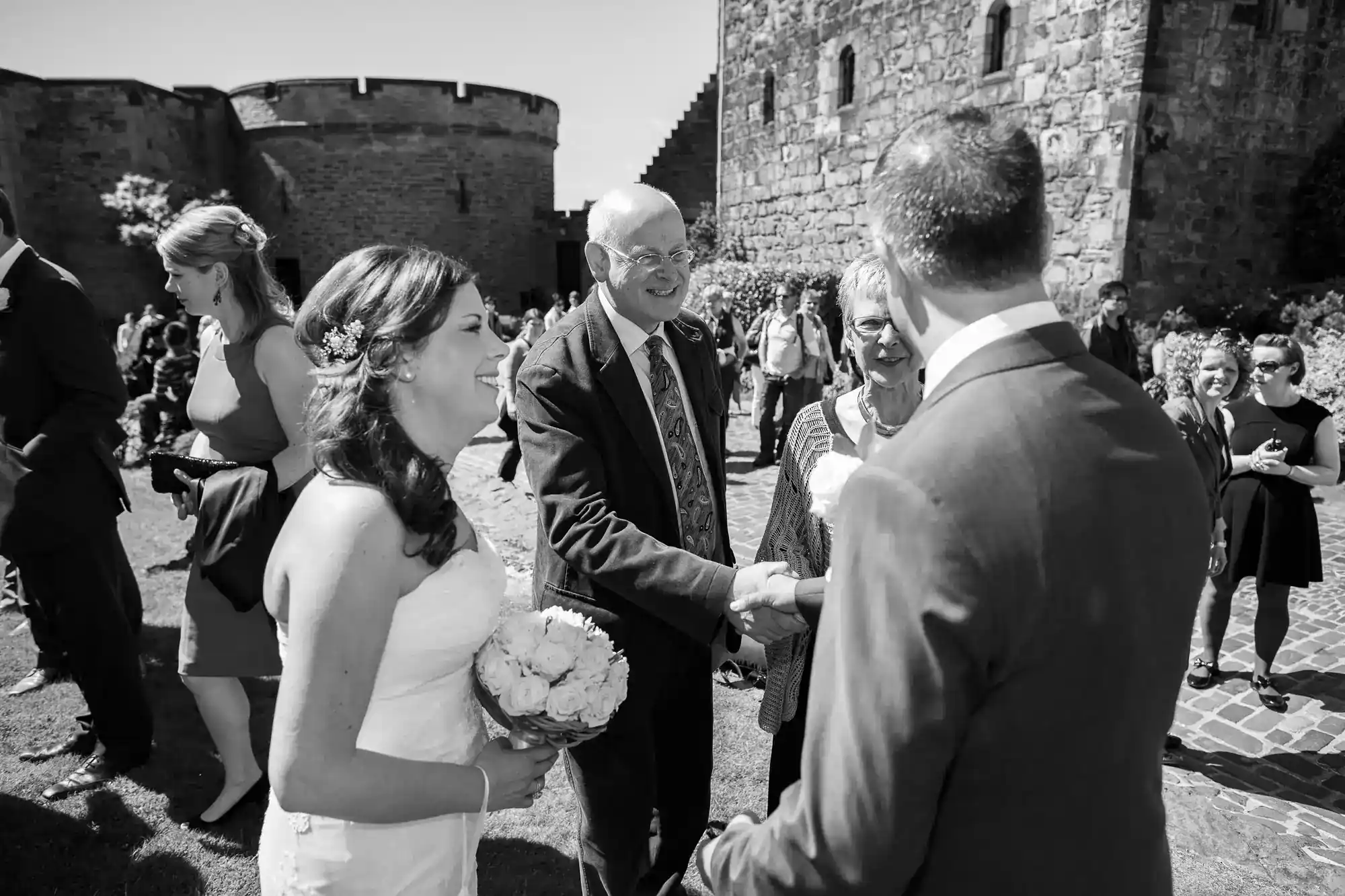A bride holding a bouquet smiles as guests greet and shake hands with a man in a suit, likely the groom, outside a stone castle. People stand and converse in the background on a sunny day.