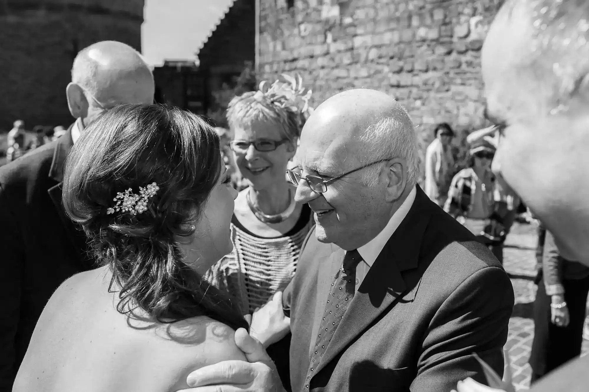 A smiling older man in a suit warmly greets a woman in a dress with a jewelled hair clip. Others, including a cheerful woman with glasses and a fascinator, look on. They are outdoors near a stone building, in bright sunlight.