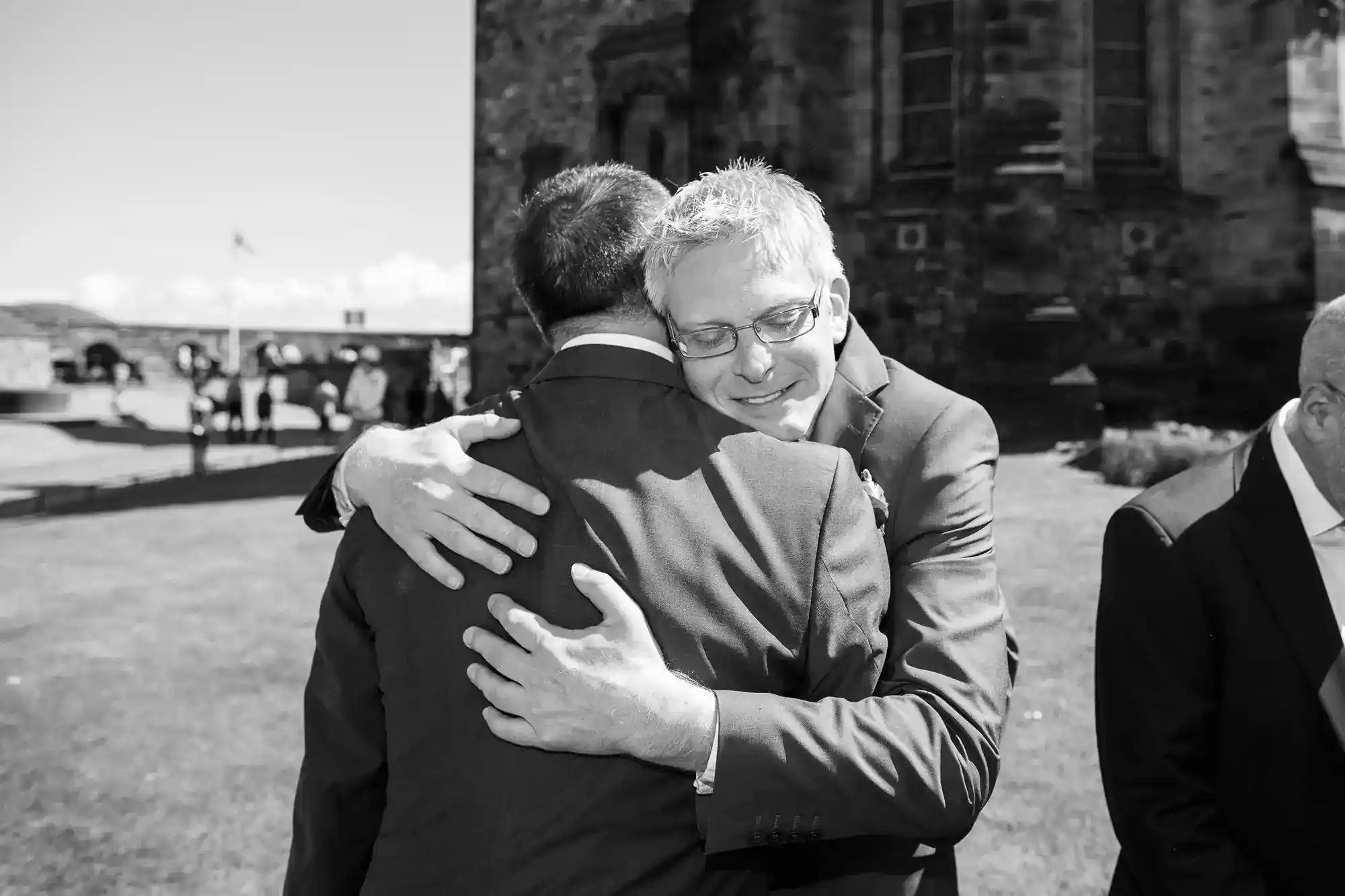 Two men in suits embrace warmly outside in sunlight, one facing the camera with eyes closed and a gentle smile. A stone building and people in the background suggest a formal or celebratory event, possibly a wedding.