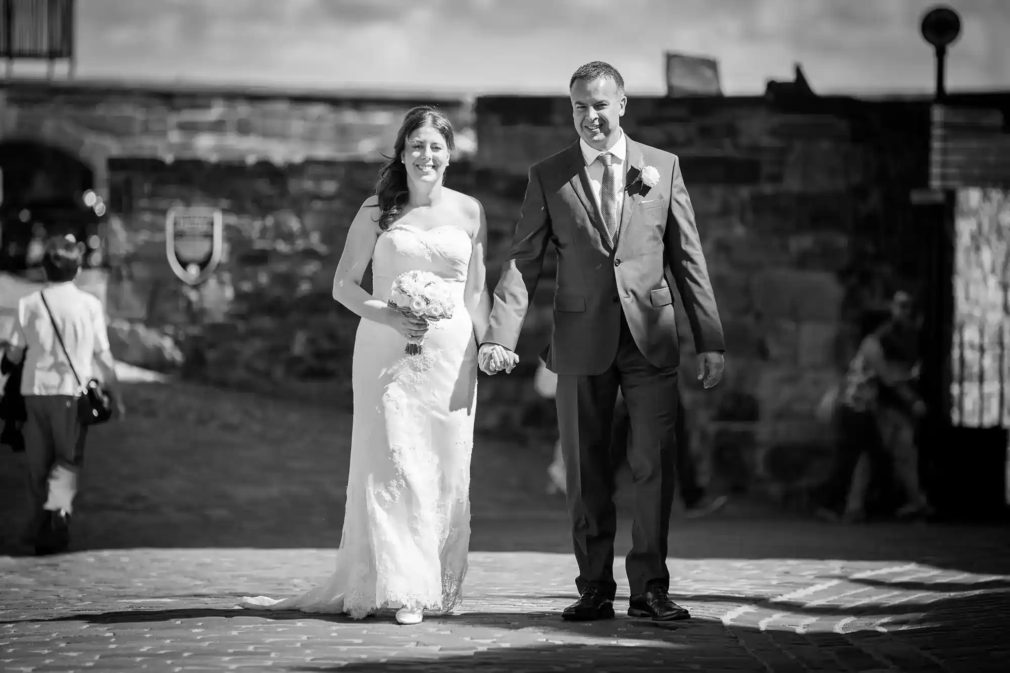 A bride in a strapless white gown holding a bouquet and a groom in a suit walk hand-in-hand outdoors, both smiling. The background shows a stone wall, other people, and bright sunlight. The photo is in black and white.