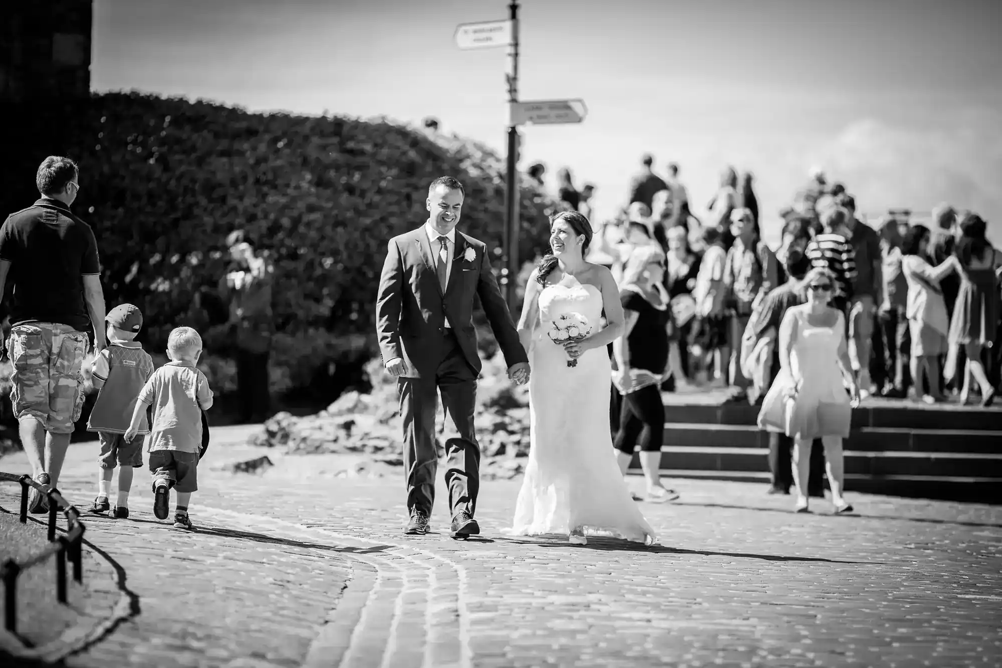 A bride and groom walk together, smiling, on a cobbled path. The groom wears a suit; the bride holds a bouquet and wears a strapless dress. People and children are in the background, near steps and a signpost, on a sunny day.