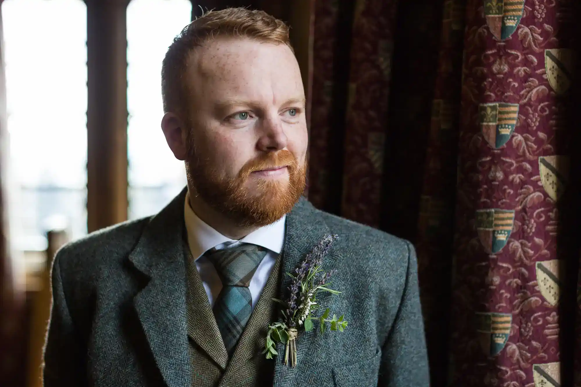 A bearded man with short red hair, wearing a grey tweed suit, tie, and buttonhole stands indoors by a window with patterned curtains, looking pensively to the side. Light from the window illuminates his face.