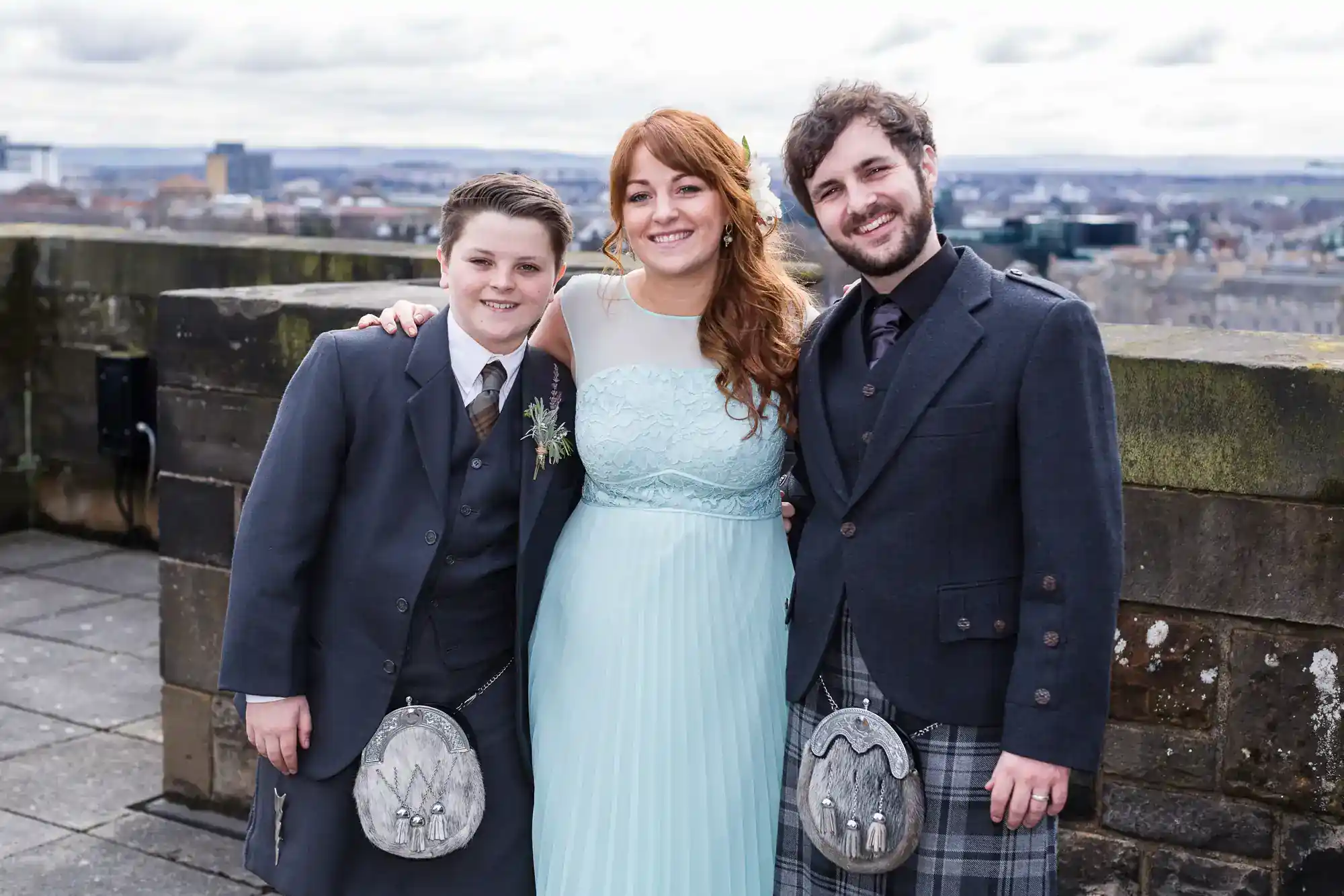 Three people smile, standing arm-in-arm outdoors. A woman in a pale blue dress is in the centre, flanked by two men in Scottish attire with kilts and sporrans. Behind them is a stone wall, and a city landscape stretches out in the background under a cloudy sky.