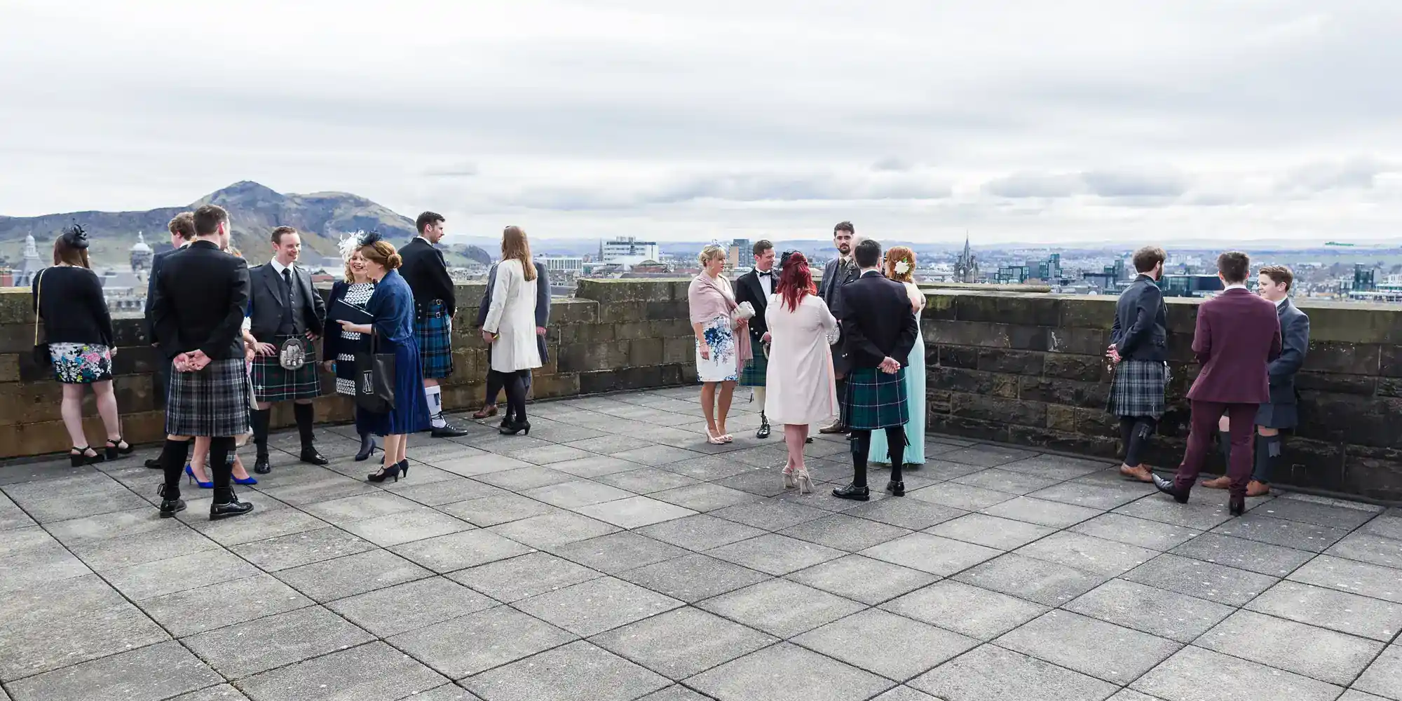A group of people, many in kilts and formal attire, stand and chat on a large stone terrace overlooking a cityscape and distant hills under a cloudy sky. The mood appears sociable and relaxed, with small groups scattered across the terrace.