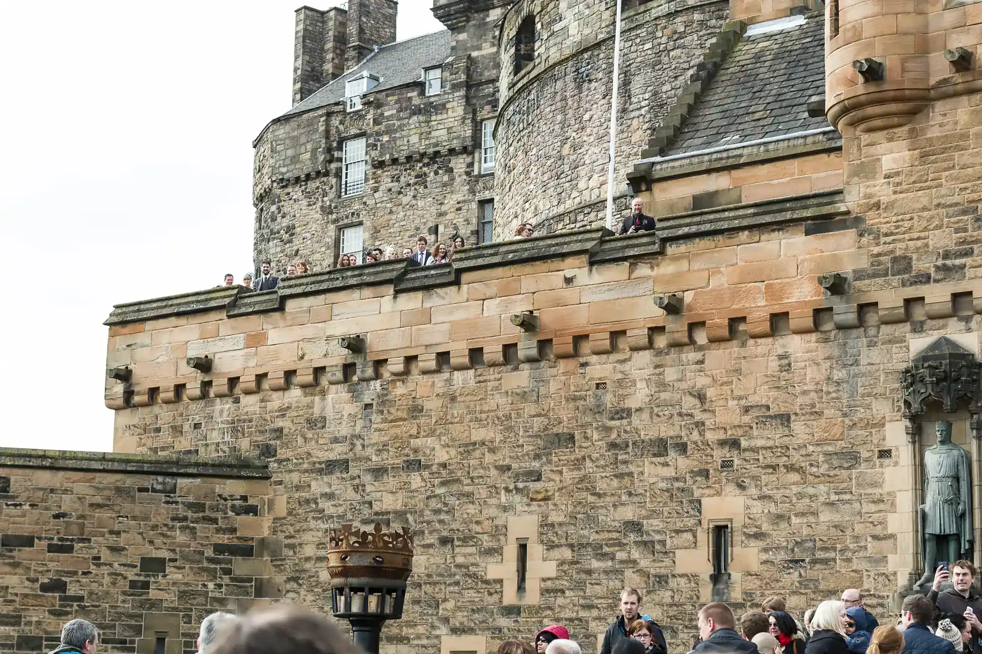 Crowds gather near a large stone castle wall, with people seen along the top and bottom. The architecture features tan bricks, small windows, and battlements. A statue and a large metal torch are visible in the lower right-hand corner.