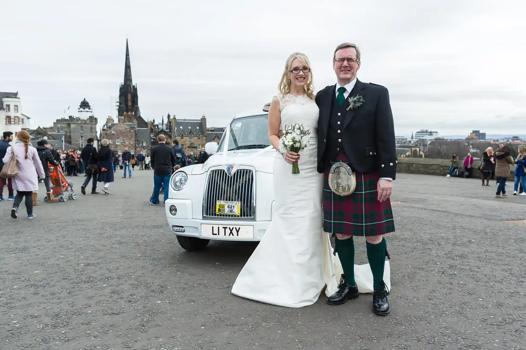 A smiling bride in a white dress stands holding a bouquet beside a groom in a kilt. They pose in front of a white car with an “L1 TXY” number plate. People and historic buildings, including a church spire, are visible in the background.
