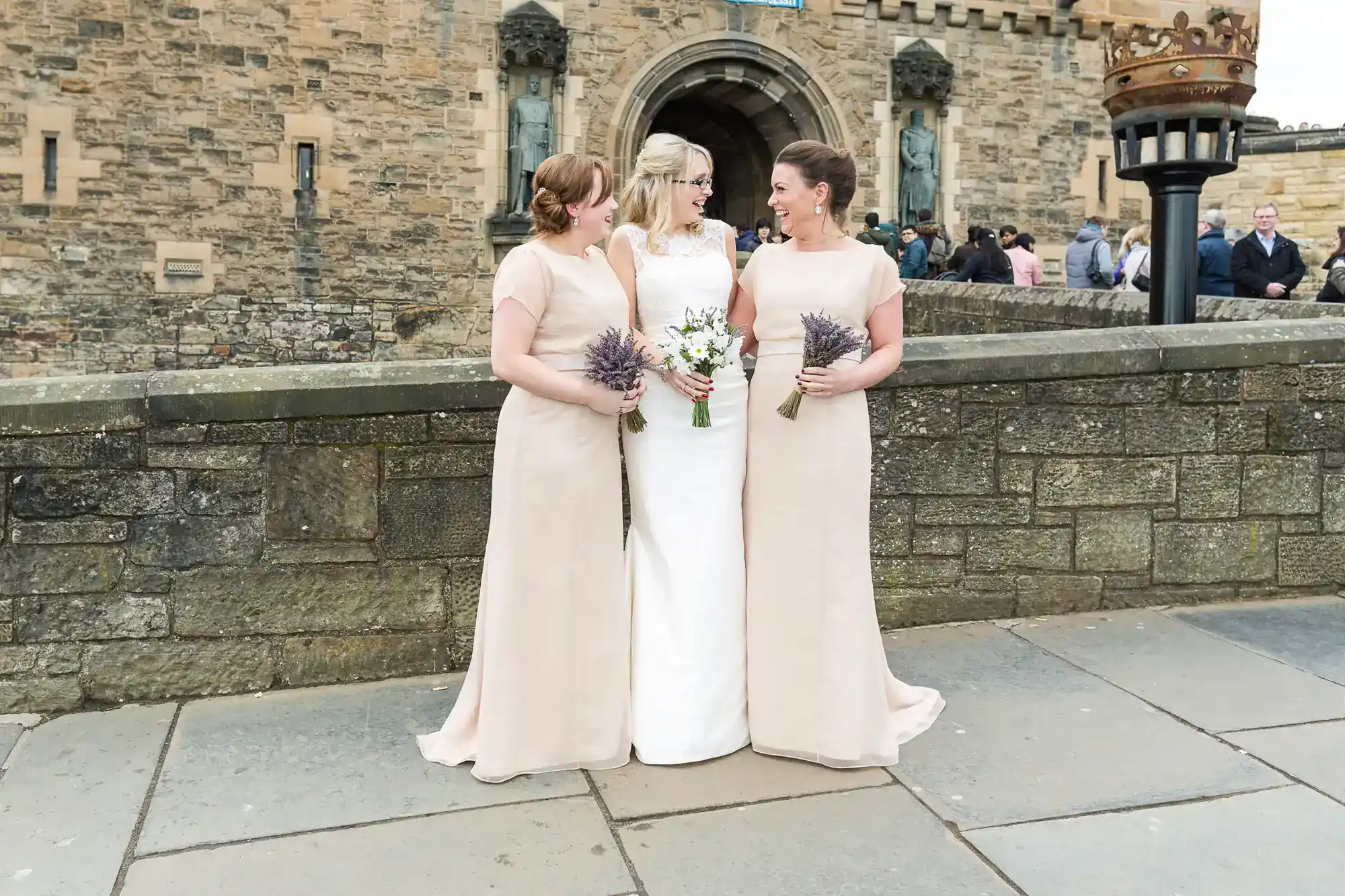 Three women in formal dresses stand on a stone walkway in front of a castle. The woman in the centre wears white and holds a bouquet, while the others wear beige dresses and hold small bouquets. They are smiling at each other.