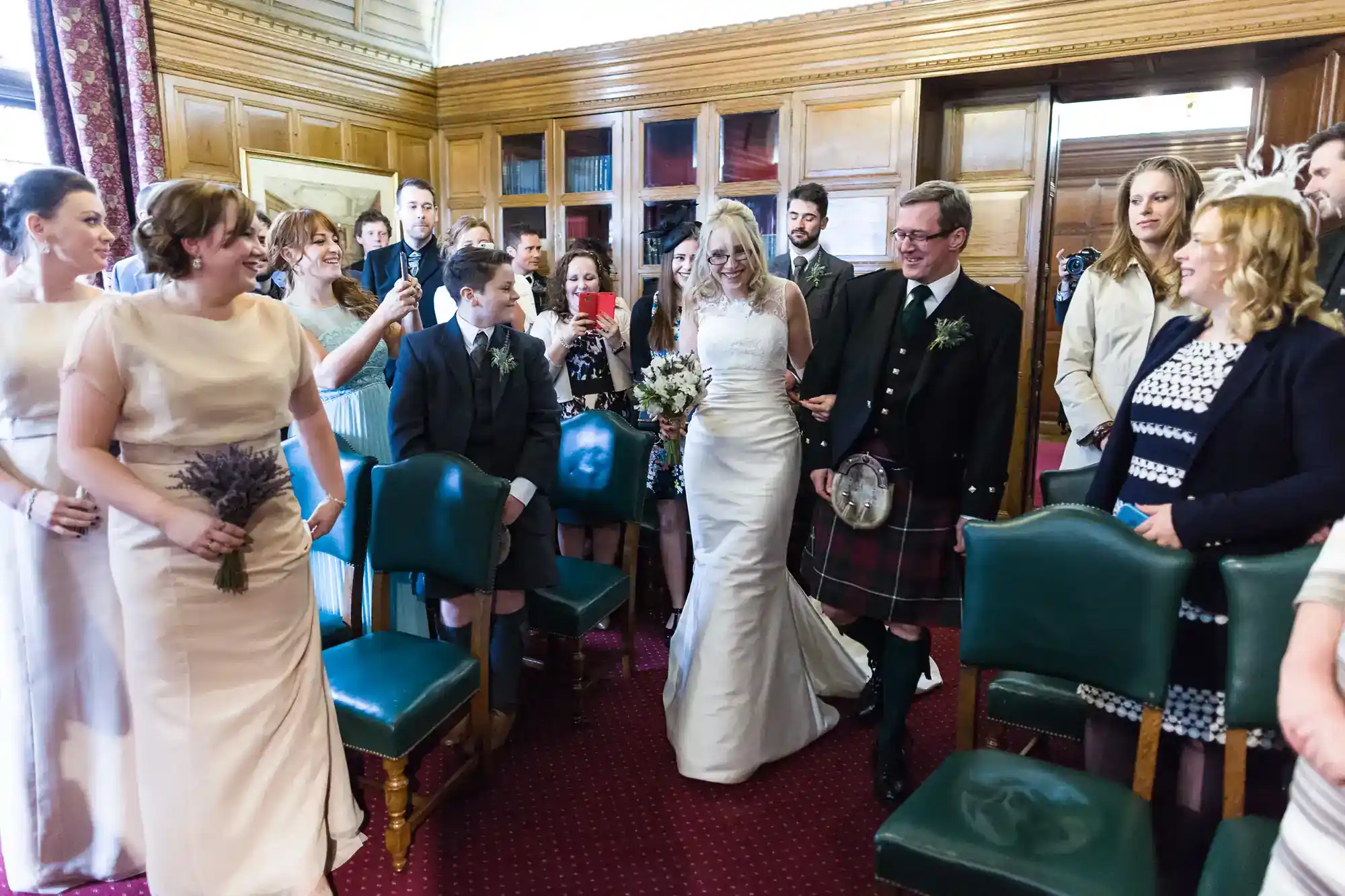 A smiling bride in a white dress walks down the aisle with a man in a kilt, surrounded by happy, well-dressed guests in a wood-panelled room. Some guests hold bouquets and take photos as they watch the bride approach.