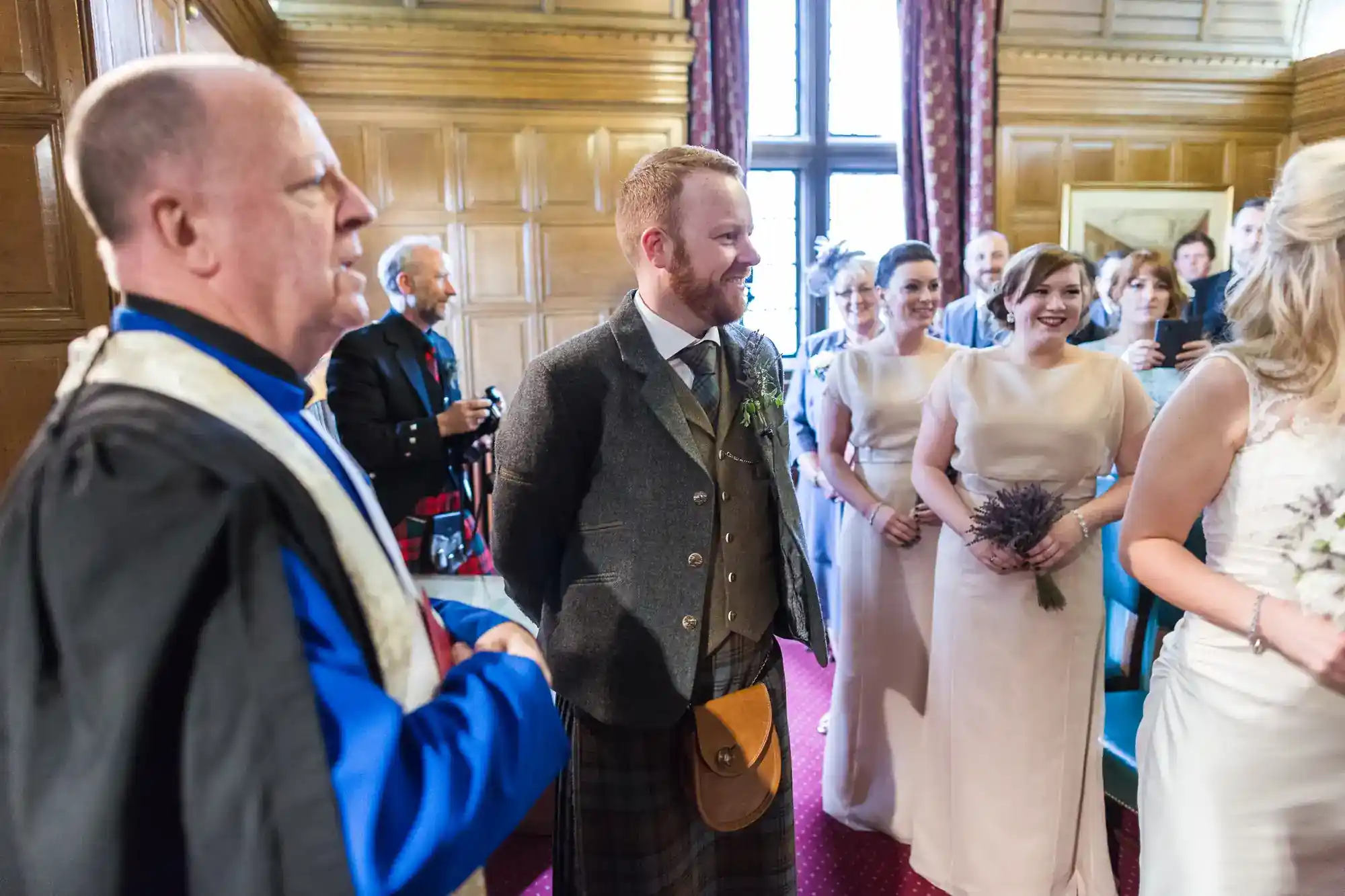A groom in a kilt stands beside an officiant in blue robes, smiling at a bride in white. Bridesmaids in blush dresses hold bouquets. Wedding guests watch, smiling, in a wood-panelled room with red carpet and large window letting in daylight.