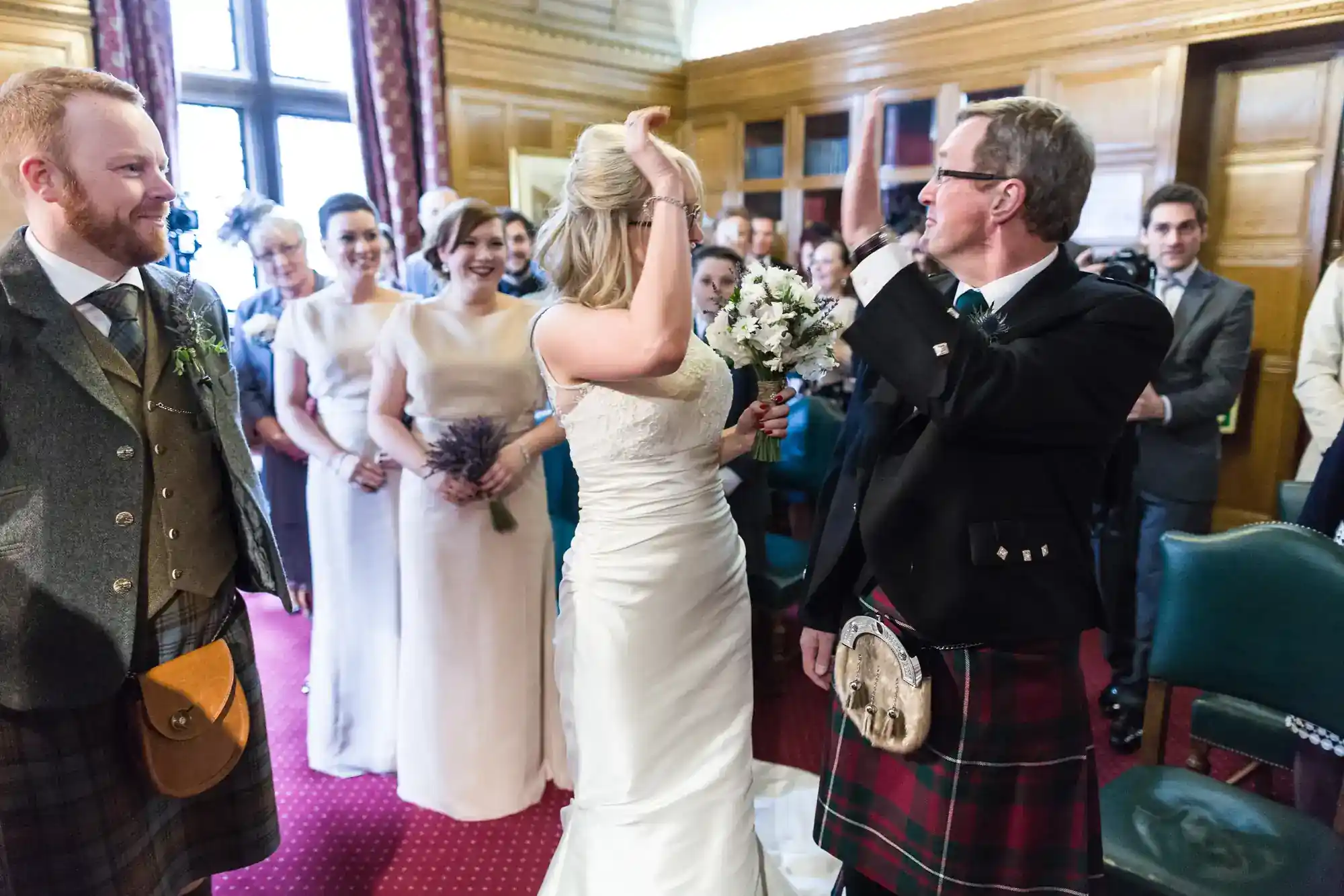 A bride in a white dress stands with a bouquet, facing an older man in a kilt. Bridesmaids in pale dresses smile behind her, and guests in suits and kilts watch in a wood-panelled room with red carpet and tall windows.