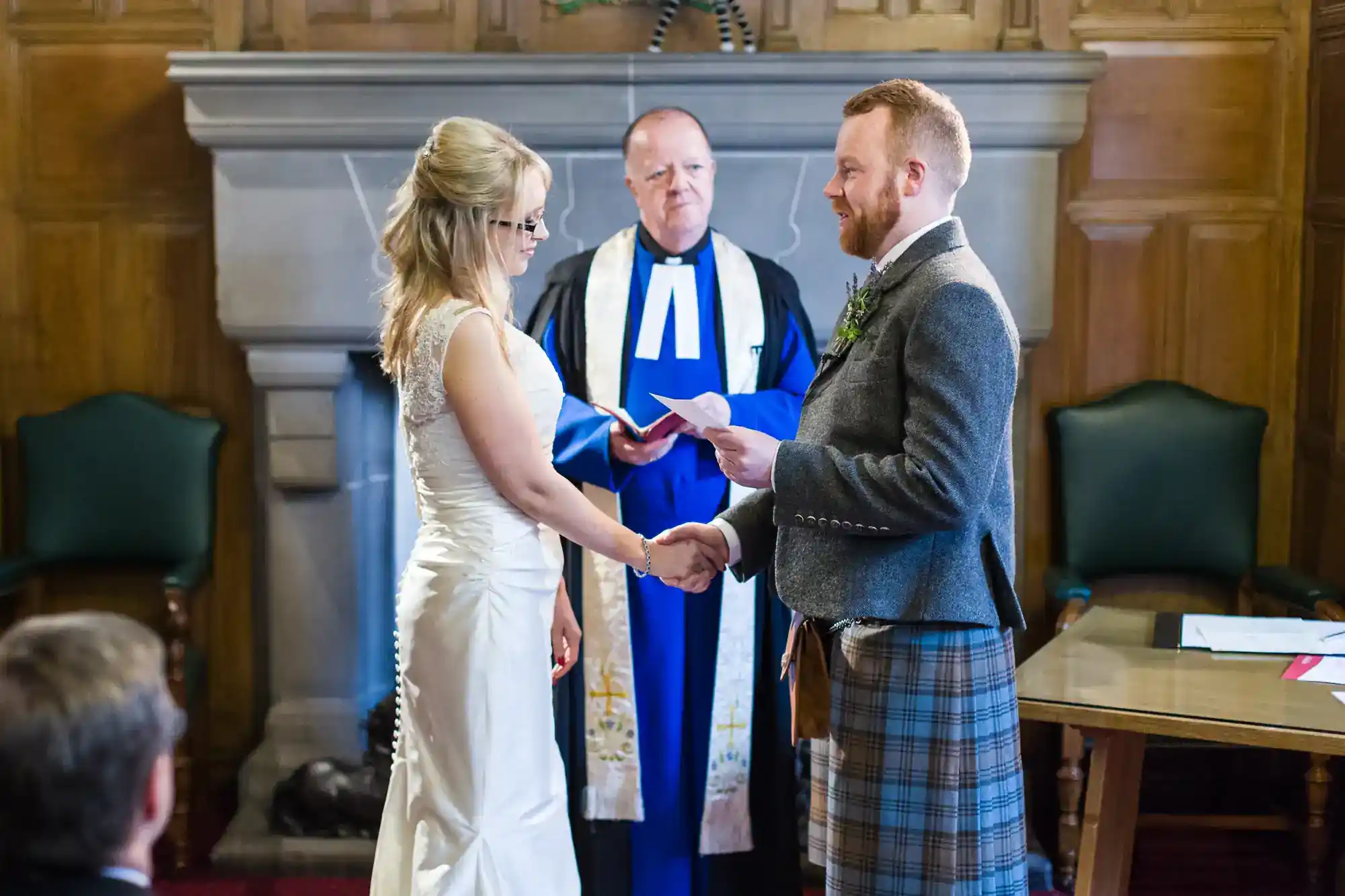 A bride in a white dress and a groom in a suit jacket and tartan kilt hold hands, facing each other, as an officiant in a blue robe stands behind them in a wood-panelled room. A table with papers and green chairs are visible nearby.