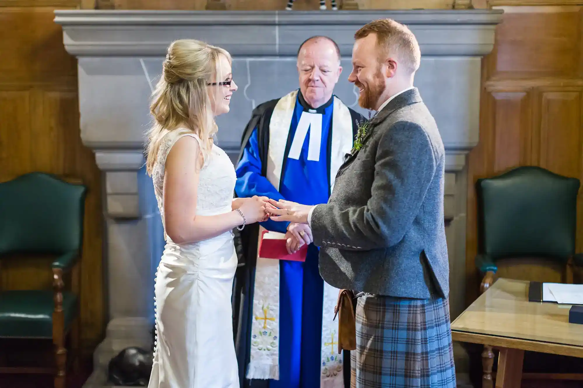 A bride in a white dress and a groom in a grey jacket and kilt exchange rings, smiling at each other, while an officiant in blue and black robes stands behind them in a wood-panelled room.