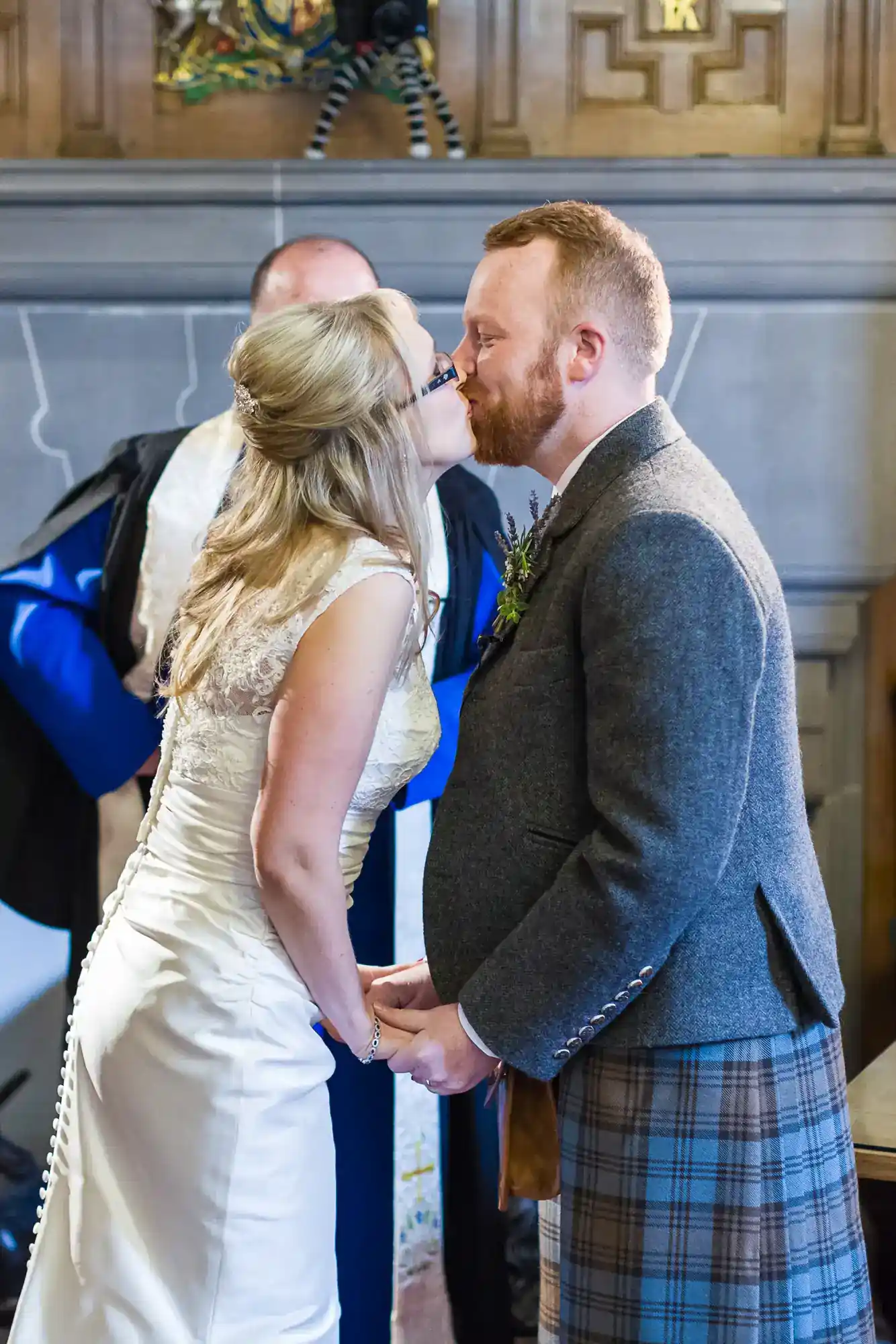 A bride and groom kiss whilst holding hands at their wedding, standing indoors. The bride wears a white, lace dress, and the groom wears a grey jacket with a checked kilt. An officiant in ceremonial robes stands behind them.