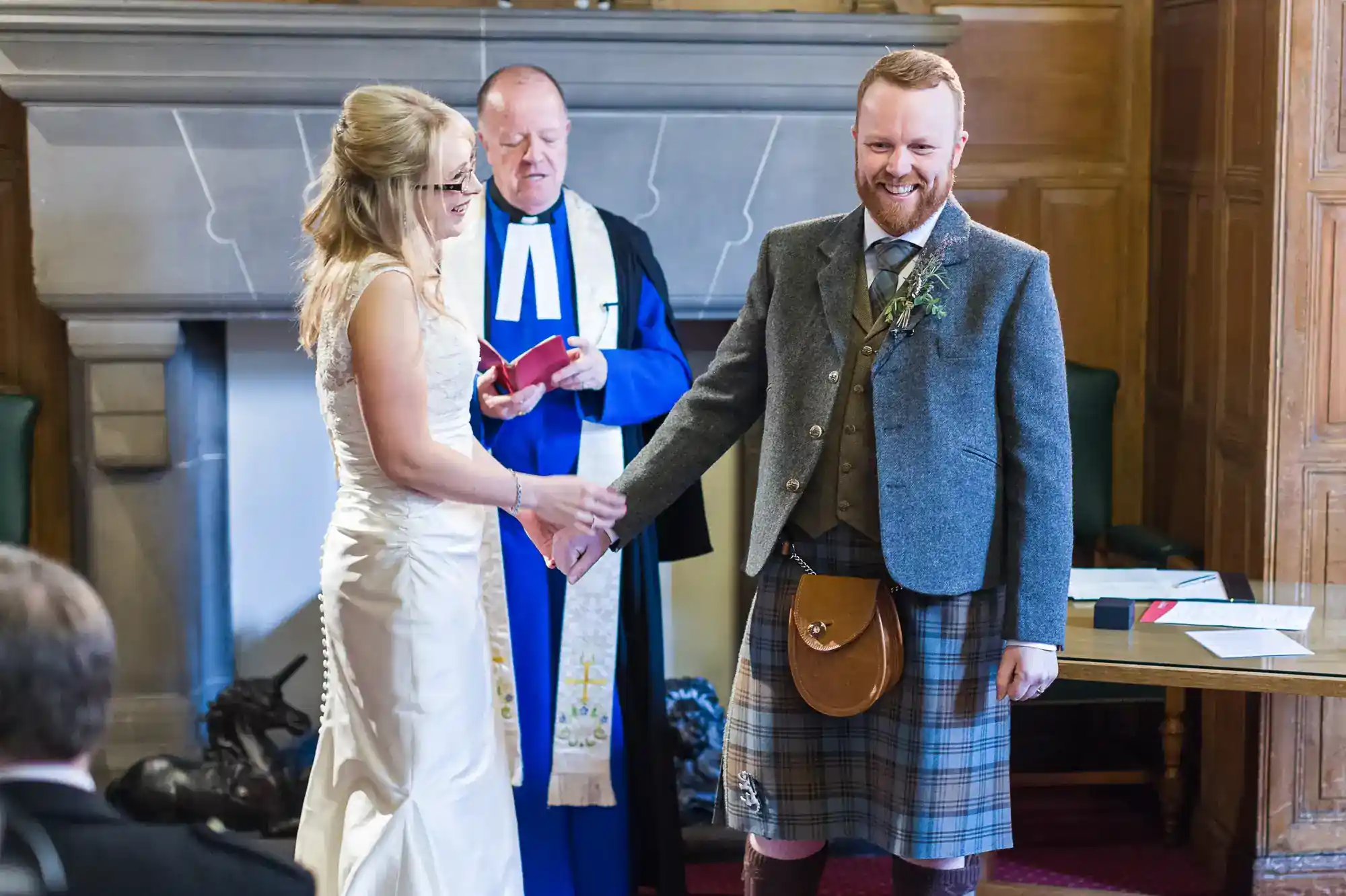 A bride in a white dress and a groom in a grey jacket and tartan kilt hold hands and smile at an indoor wedding ceremony. A minister in blue and white robes stands behind them, holding a book. The room has wood-panelled walls and a fireplace.