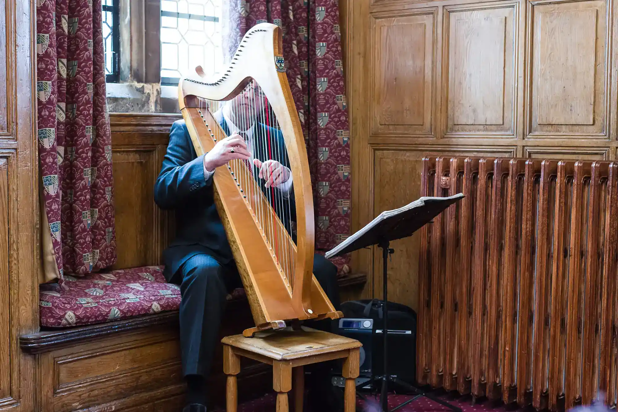 A person in a suit plays a wooden harp on a cushioned bench near a radiator in a wood-panelled room. Sheet music rests on a stand beside them. The player’s face is partly hidden by the harp, and a window is in the background.