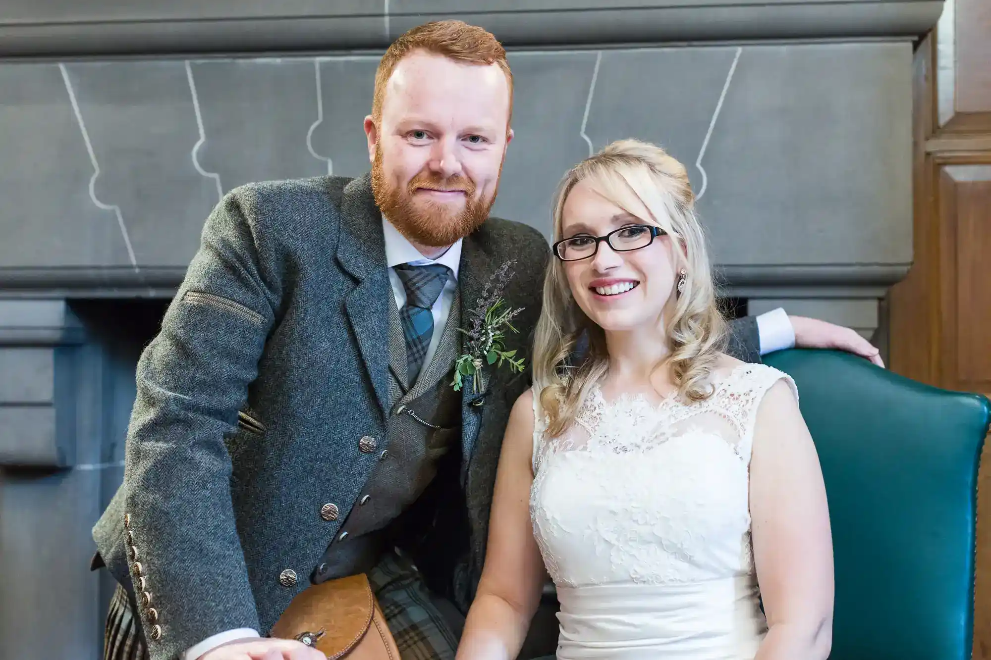A smiling man in a grey suit with a tartan kilt stands beside a seated woman in a white lace wedding dress. Both appear happy, posing in front of a grey fireplace, with the man’s arm resting on the chair.