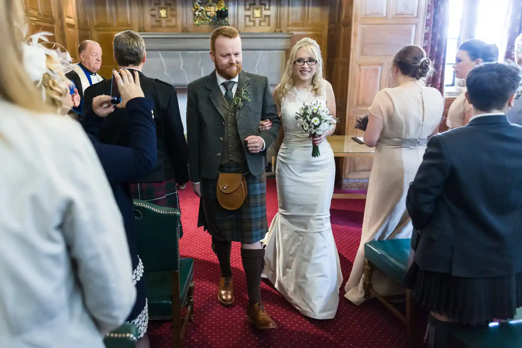A bride in a white gown and glasses walks arm-in-arm with a groom in a kilt and tweed jacket down an aisle, smiling. Guests in formal attire, some in kilts, watch and take photos in a wood-panelled room with red carpet and a fireplace.