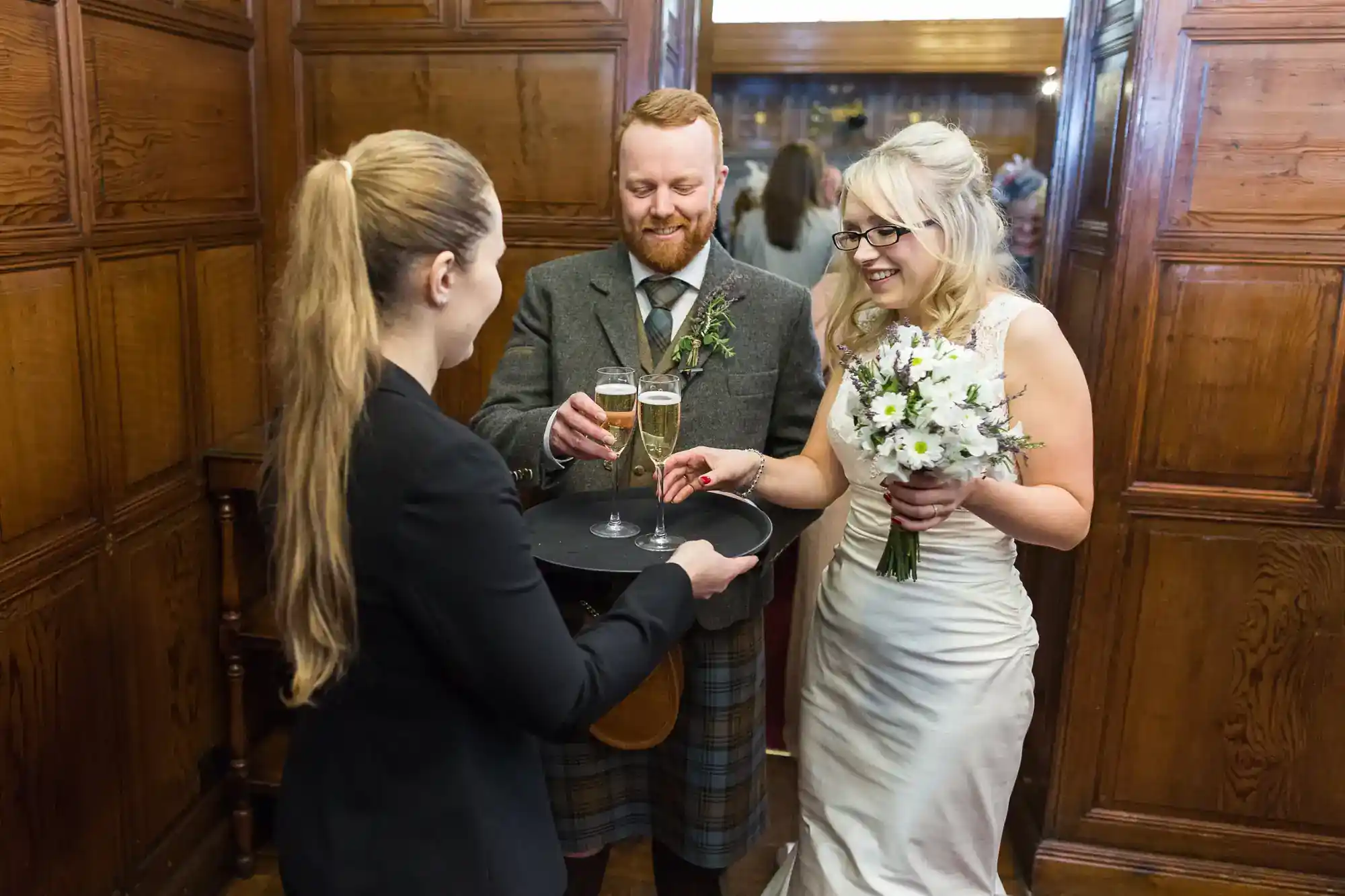A smiling bride in a white dress holds a bouquet as a waiter hands champagne to a groom in a kilt. They stand in a wood-panelled room with a mirror, reflecting guests and adding depth to the celebratory wedding moment.