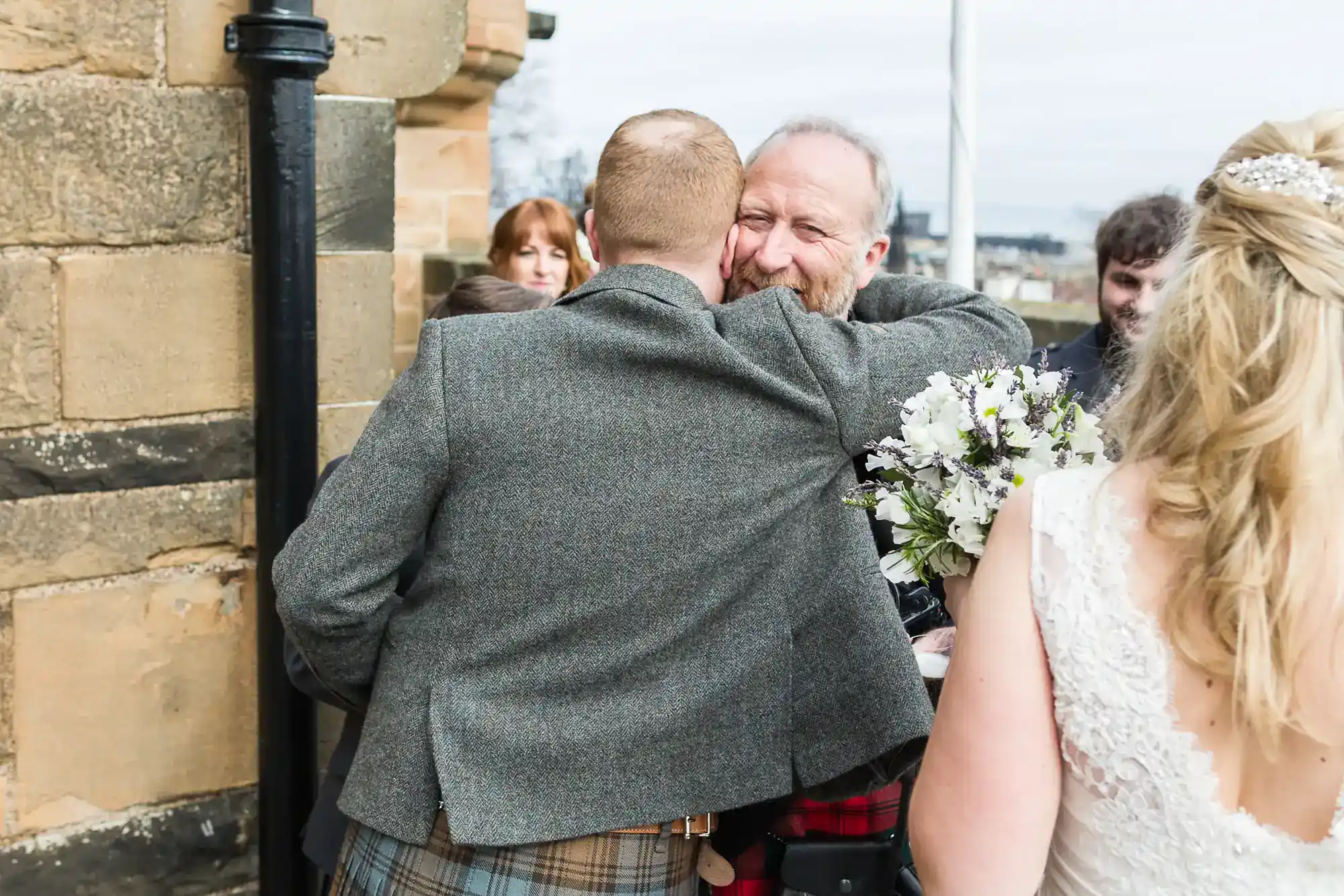 Two men in tweed embrace joyfully outside a stone building. One man holds a bouquet. A woman in a white lace dress stands with her back to the camera. Several people watch in the background. It appears to be a wedding celebration.