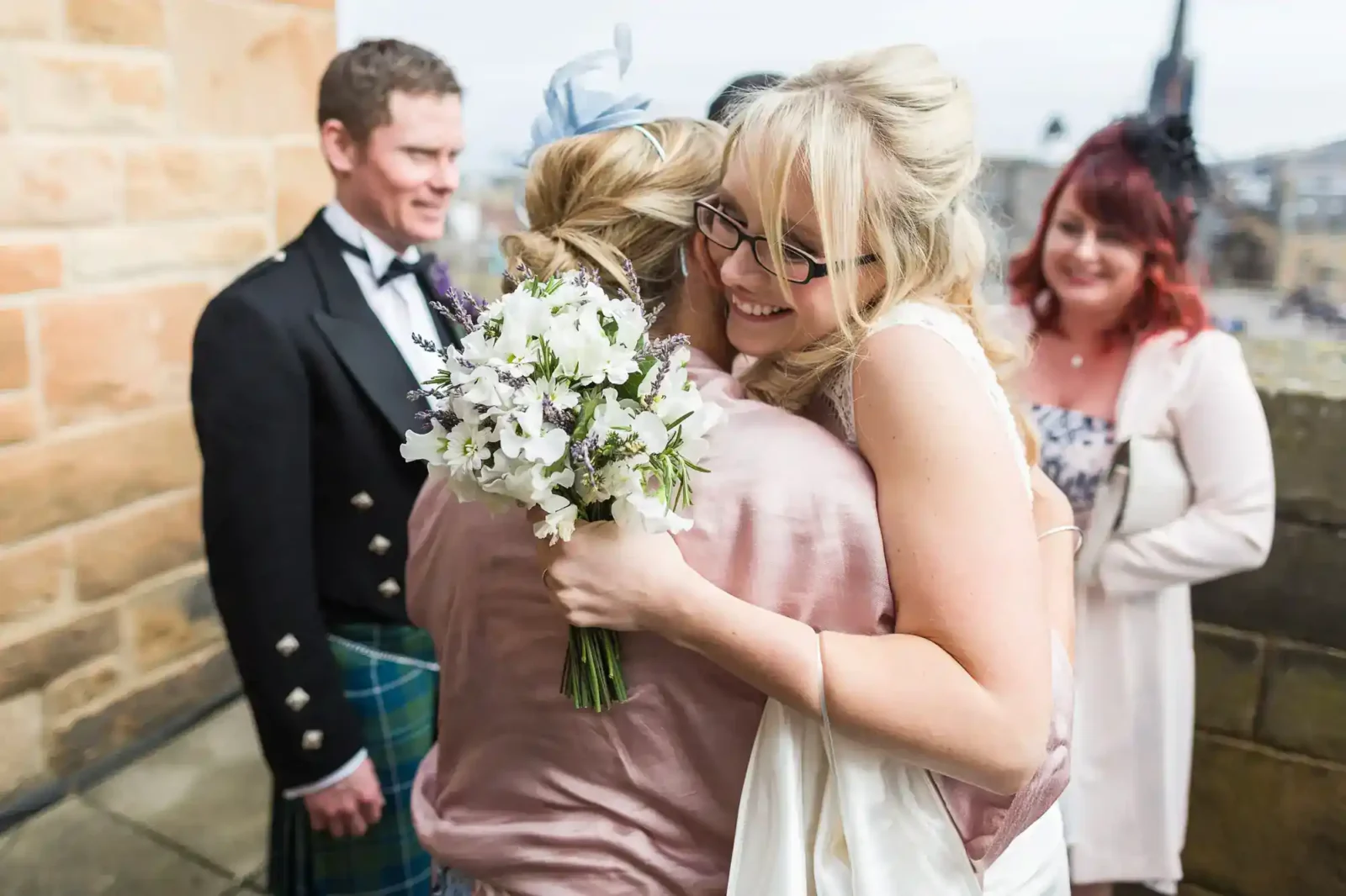 A smiling bride in glasses hugs a guest holding a bouquet of white and purple flowers. Two other guests, one in a kilt and another in a floral dress, stand nearby, smiling. The scene is outdoors beside a stone wall.