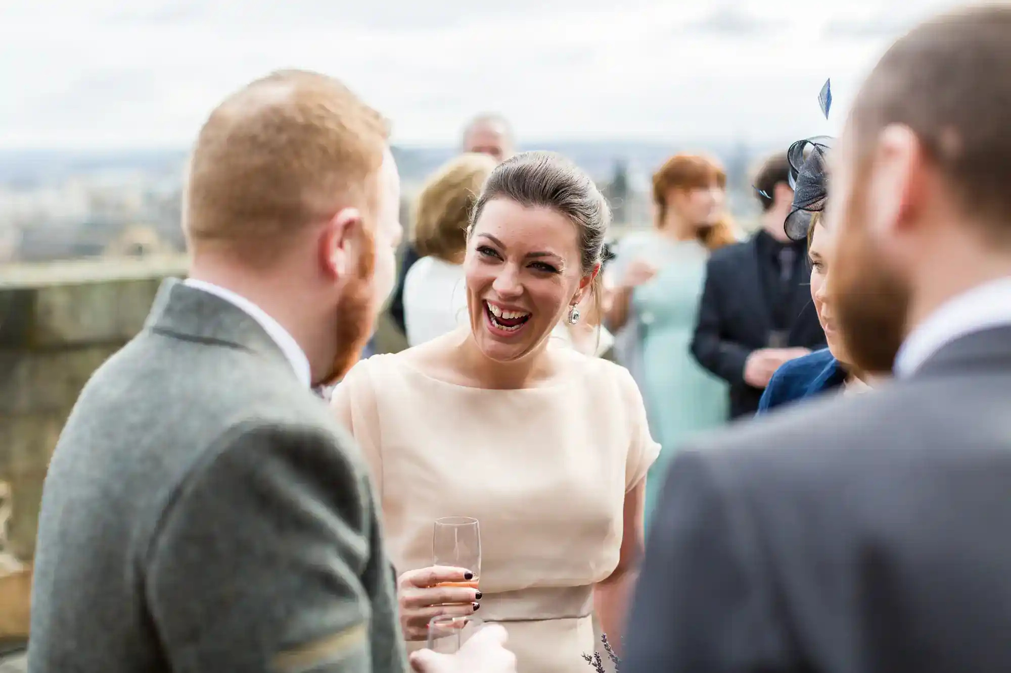 A woman in a light dress laughs while holding a drink, surrounded by people in formal attire at an outdoor event. The background is blurred, suggesting an elevated, scenic location. The mood is joyful and sociable.
