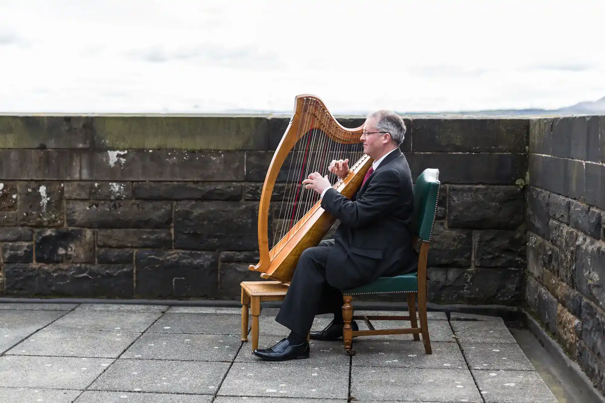 A man in a suit sits on a green chair outdoors, playing a large wooden harp beside a stone wall. The sky is overcast, and the ground is paved with grey tiles.