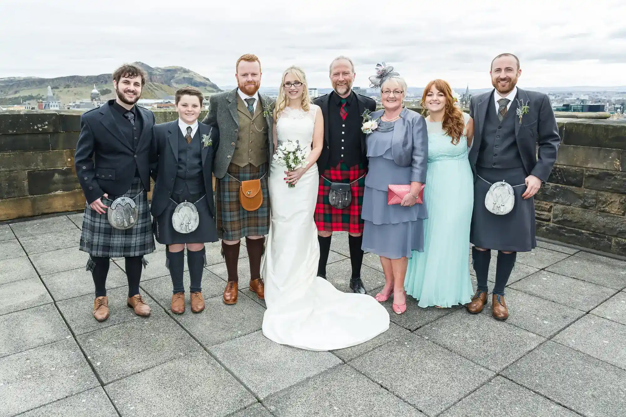 Eight people pose on a rooftop. Four men and a boy wear kilts. A bride in a white dress stands centre beside a man in a red tartan kilt. Two women in light blue dresses hold clutch bags. Everyone smiles at the camera. A hilly landscape appears in the background.