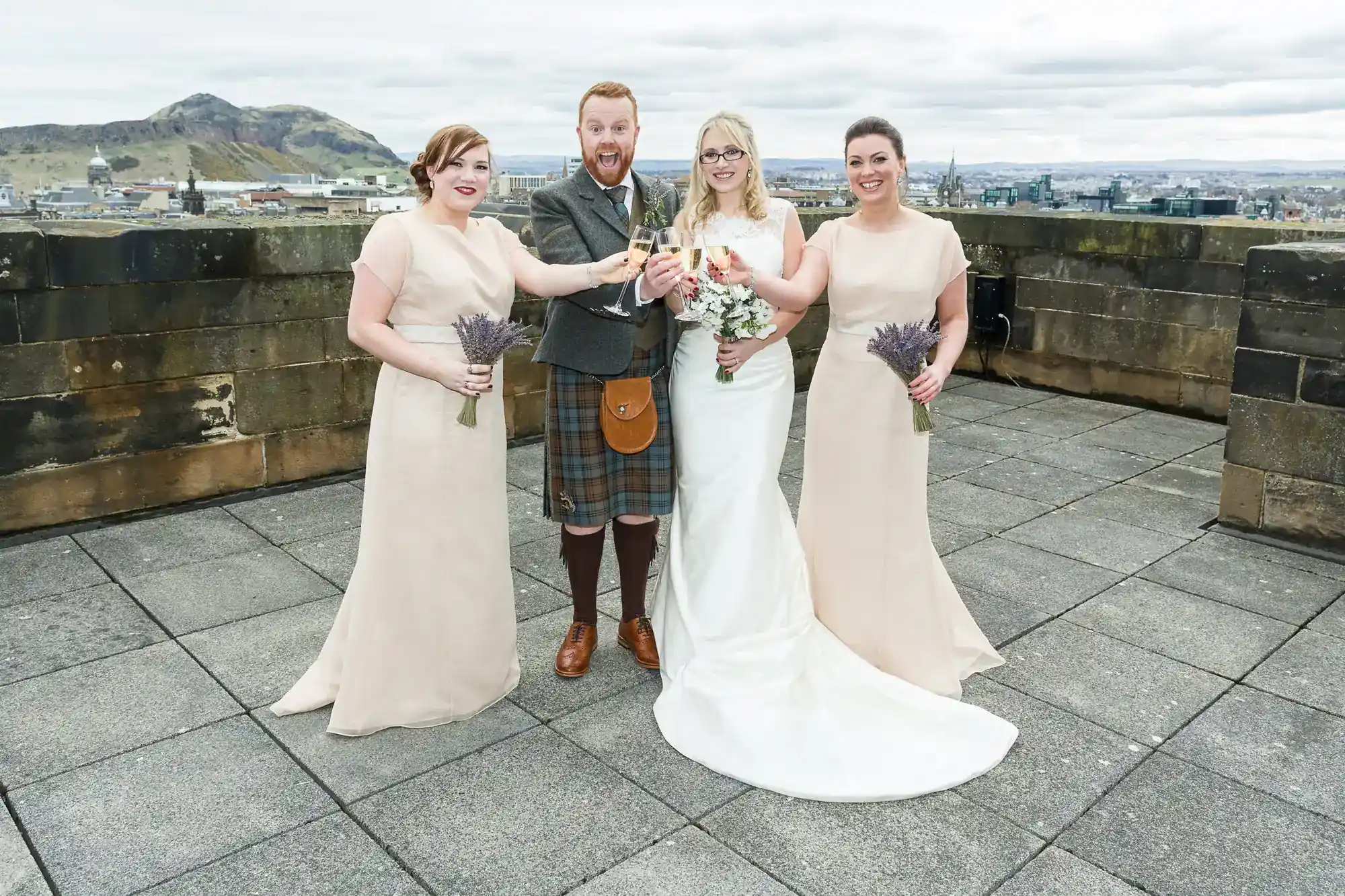 Four people pose on a rooftop: a man in a kilt holds a drink, flanked by three women in light dresses holding bouquets. The woman next to him wears a long white gown, holding a bouquet of pink and white flowers. A cityscape and hills are visible behind them.