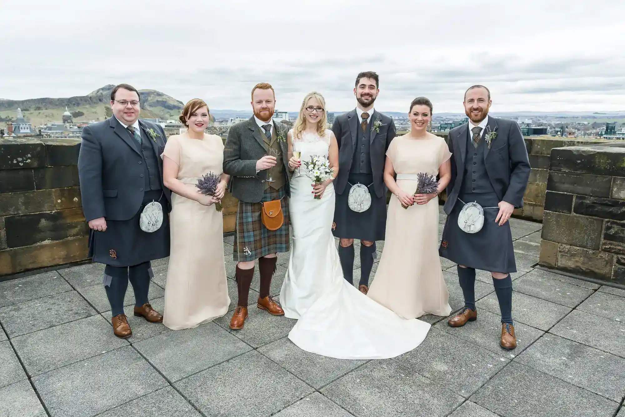 Seven people pose outdoors on a stone terrace. Three men wear kilts and jackets, two women wear light dresses and hold bouquets, the bride wears a white gown and glasses, and the groom stands beside her. Hills and cloudy sky are in the background.