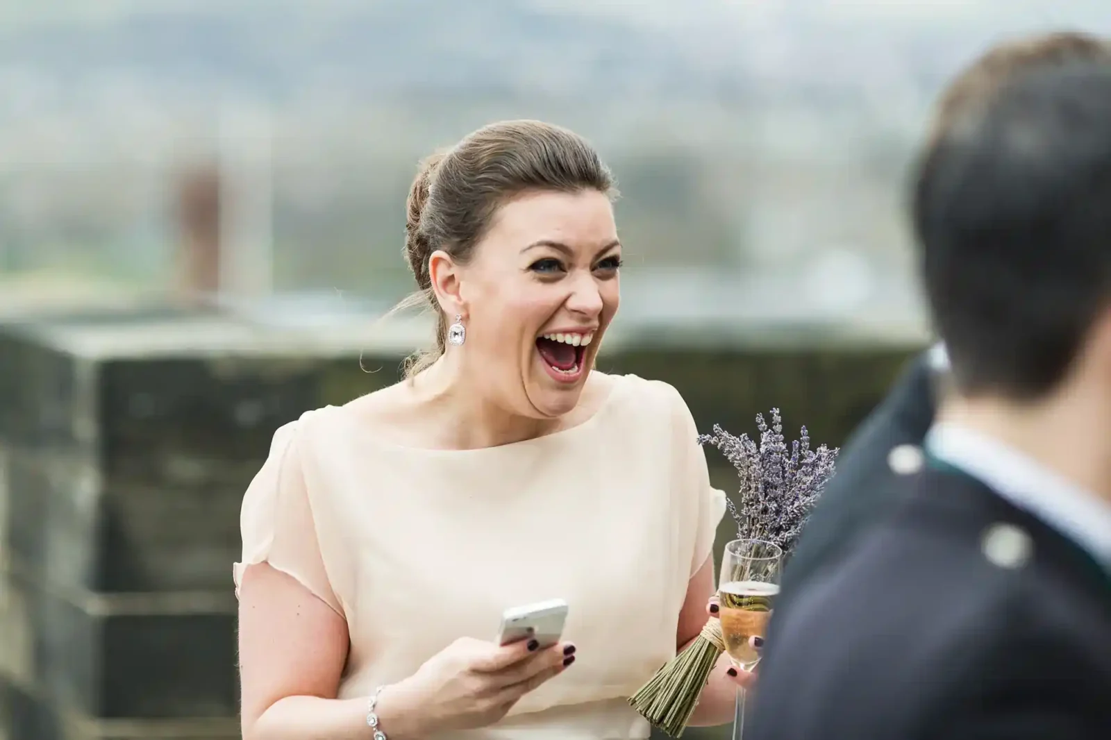 A woman in a light beige dress laughs joyfully, holding a mobile, a bouquet of lavender, and a drink. She has her hair styled up and is wearing earrings. The background is blurred, with another person partially visible in the foreground.