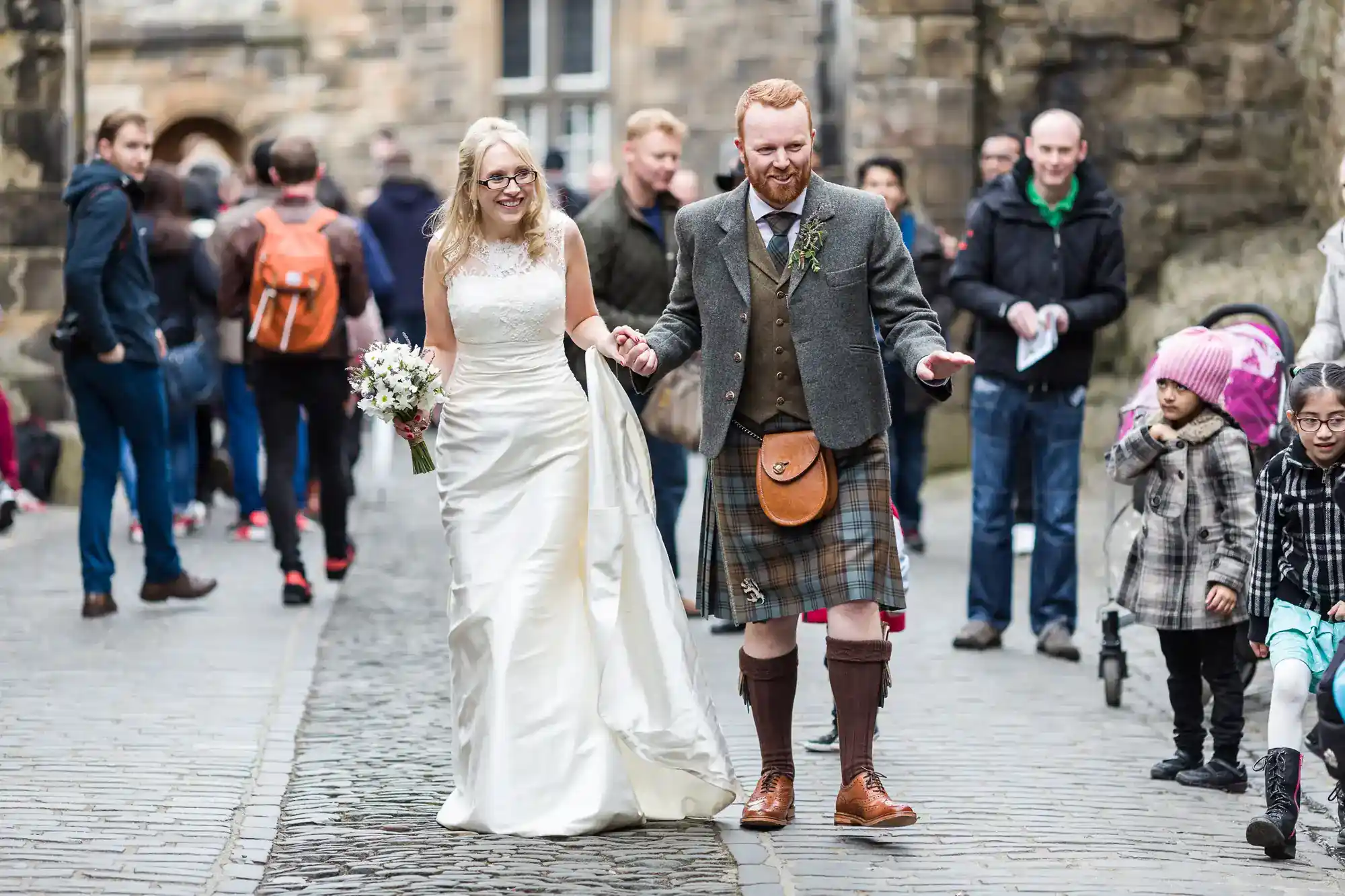 A smiling bride in a white dress and a groom in a kilt walk hand-in-hand on a cobbled street. People in casual clothes walk around them. The bride holds a bouquet, and the couple appear happy and relaxed in a lively outdoor setting.