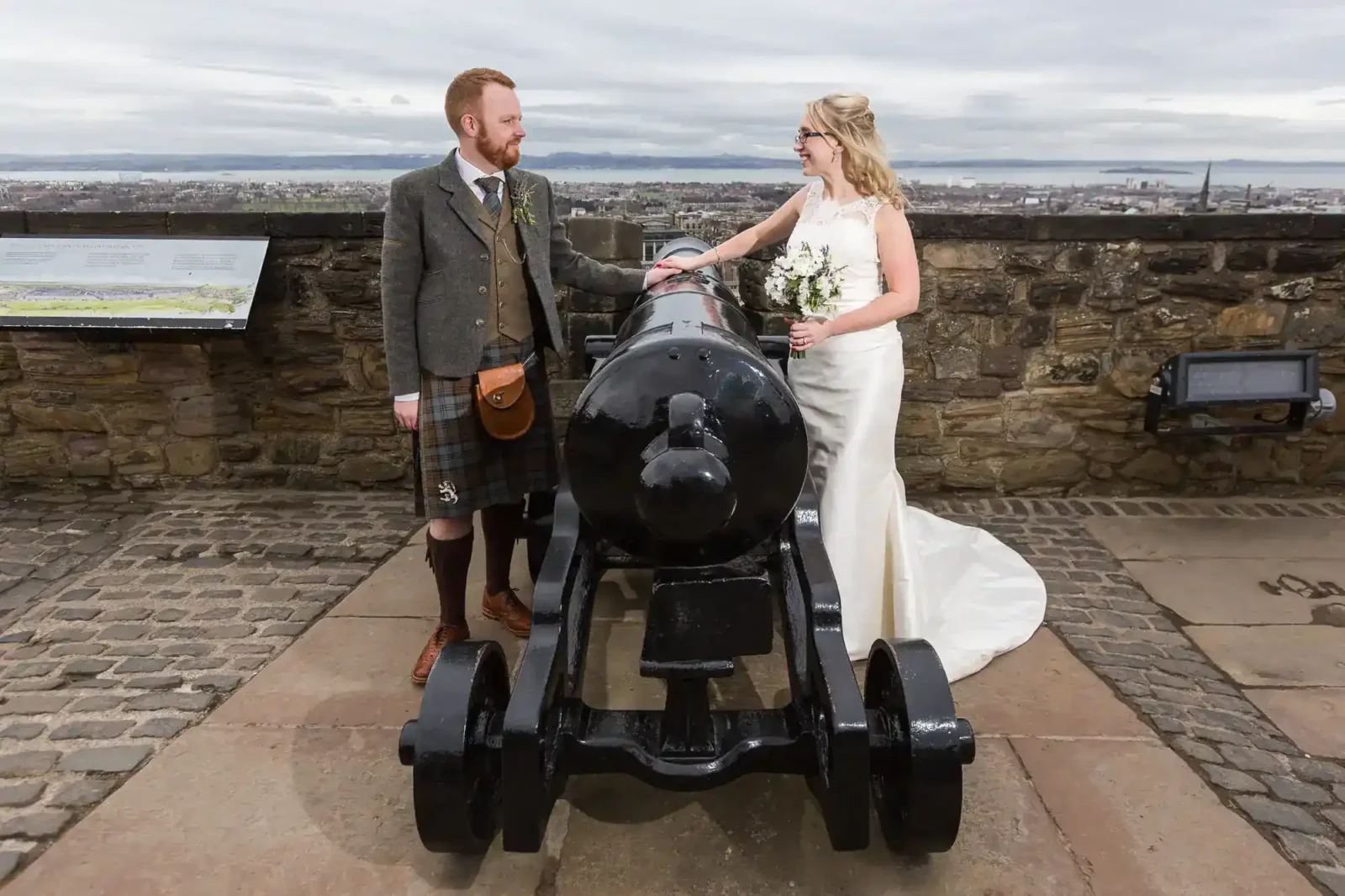 A bride in a white dress and a groom in Scottish attire with a kilt stand on either side of a large black cannon at The Castle Ramparts at Edinburgh Castle. The bride holds a bouquet; both smile at each other. Edinburgh's cityscape is visible in the background under a cloudy sky.