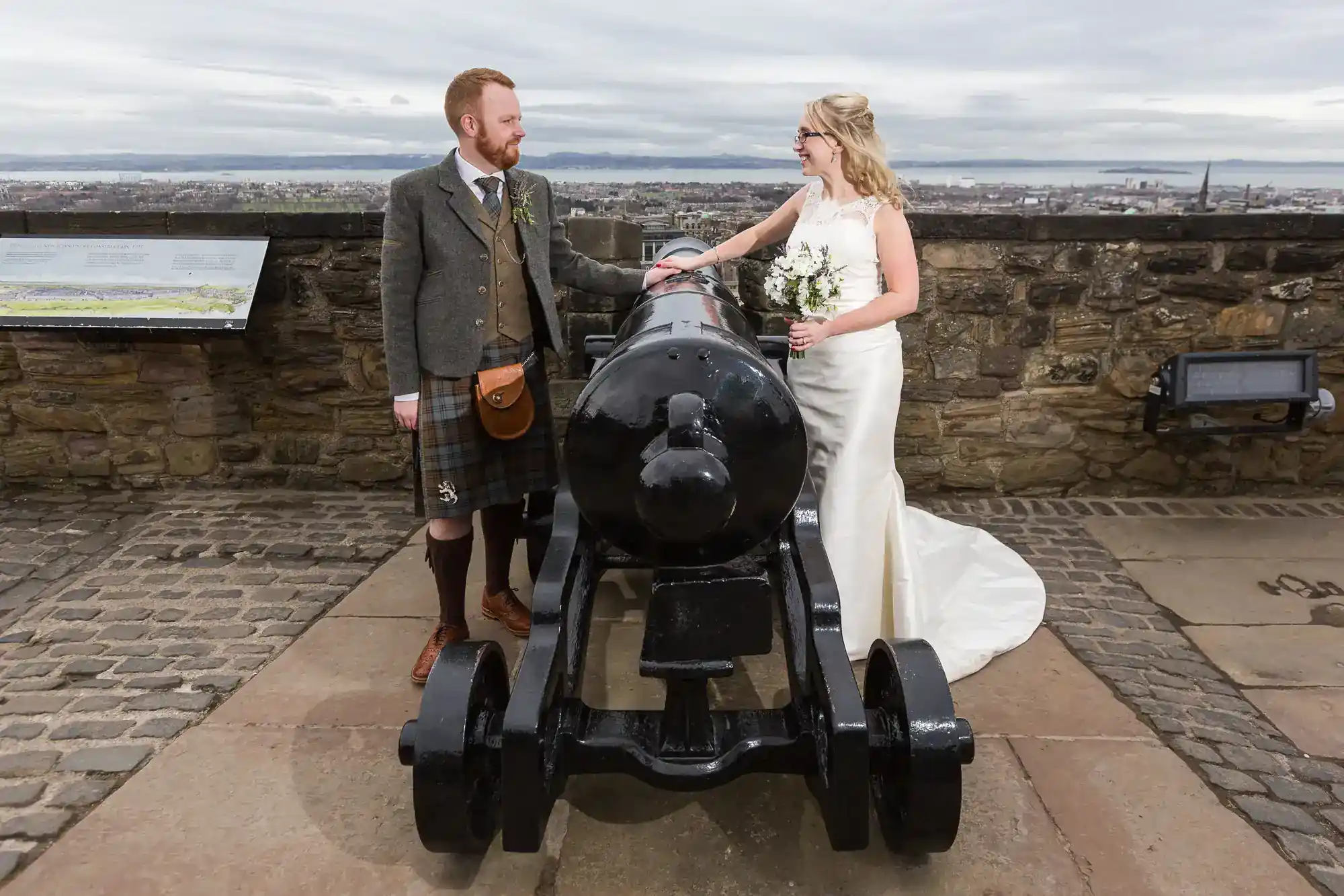 A bride in a white dress and a groom in Scottish attire with a kilt stand on either side of a large black cannon at a stone fort. The bride holds a bouquet; both smile at each other. Cityscape is visible in the background under a cloudy sky.