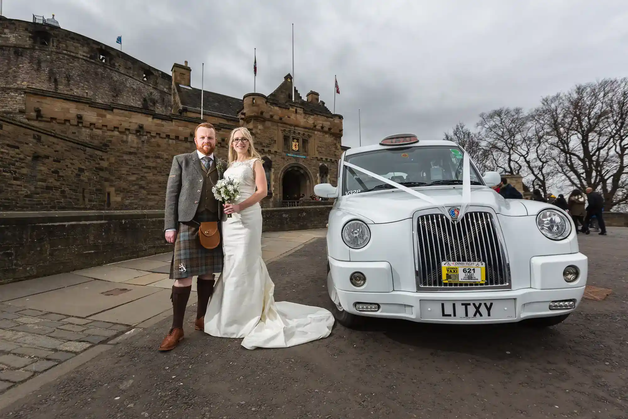 A bride in a white dress and a groom in a kilt stand smiling beside a decorated white taxi outside a historic stone castle on a cloudy day. The car is adorned with ribbons, and the castle features flags and battlements in the background.