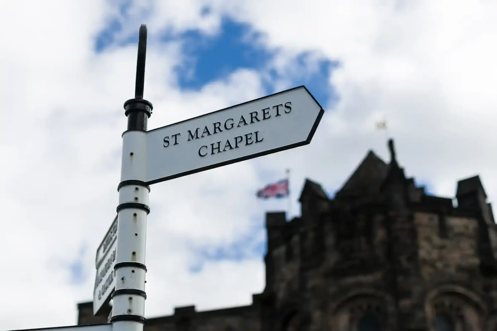 A white directional sign reading “St Margarets Chapel” points to the right. In the blurred background is a stone building with a British flag flying on top, and the sky is partly cloudy.