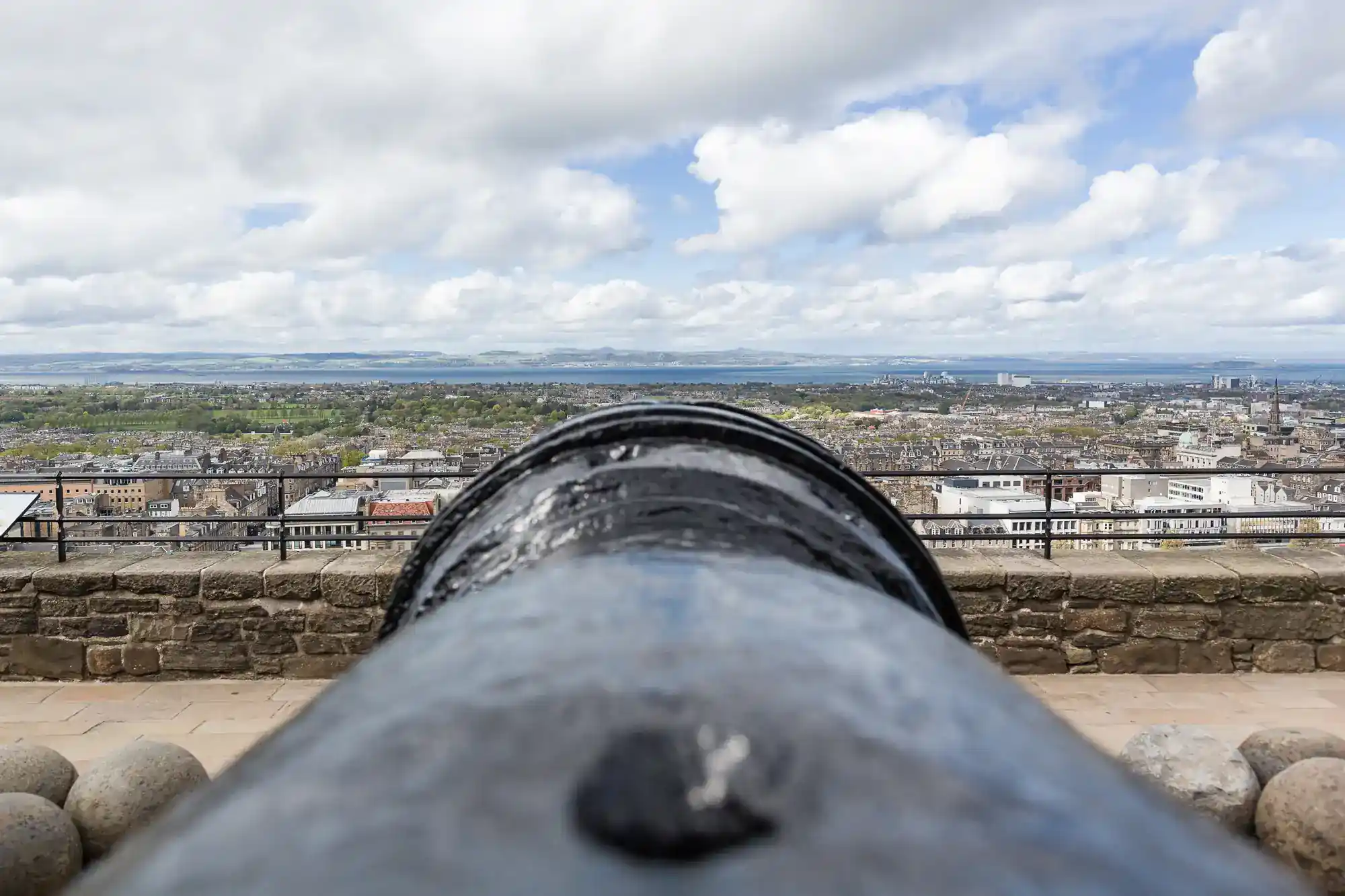 A black cannon points over a stone wall towards a cityscape under a partly cloudy sky. The foreground is dominated by the cannon, whilst buildings, greenery, and distant hills are visible in the background.